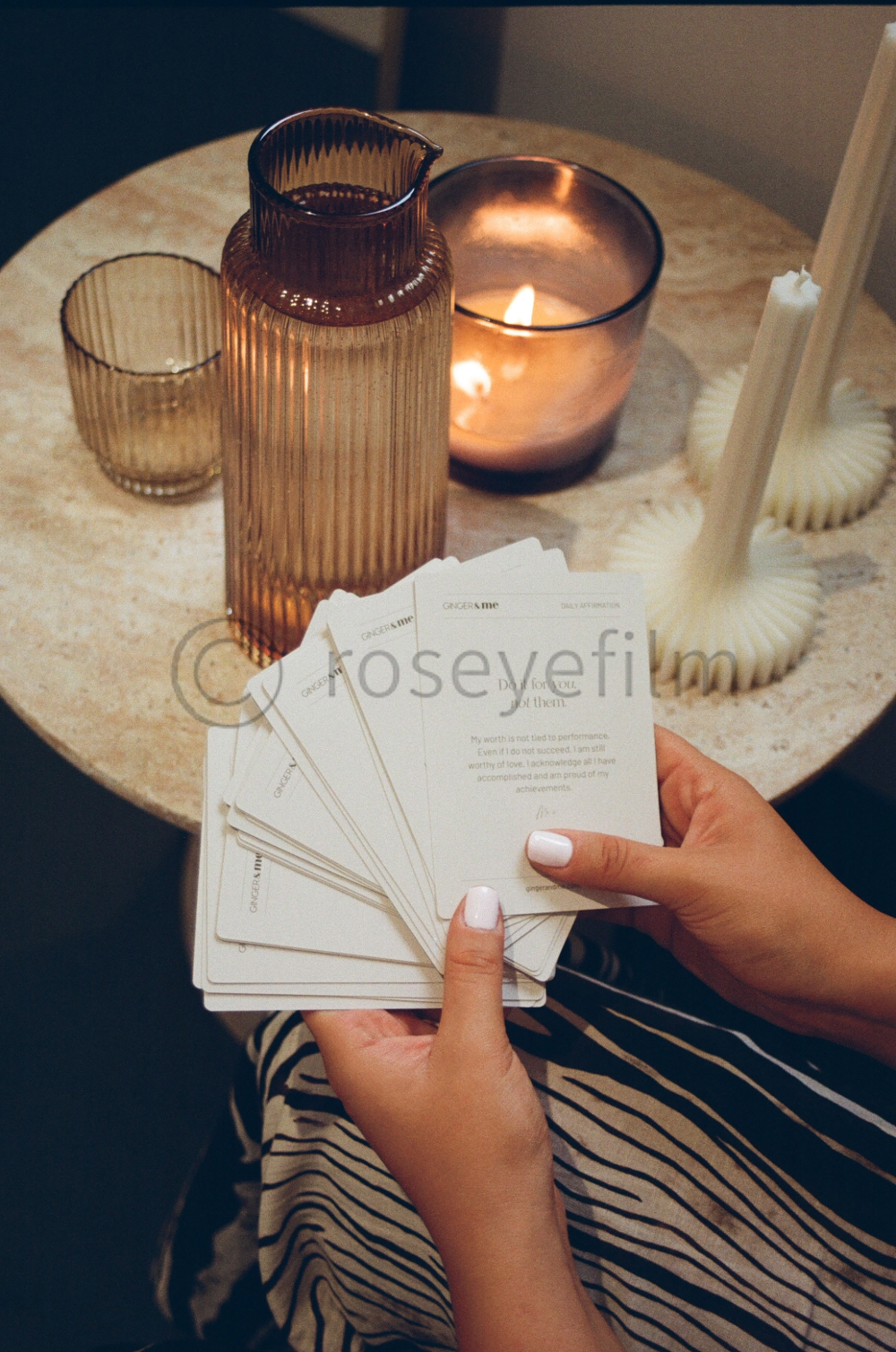 A person holding a deck of cards with inspirational messages, sitting at a table with candles, a decorative vase, and a small drinking glass.