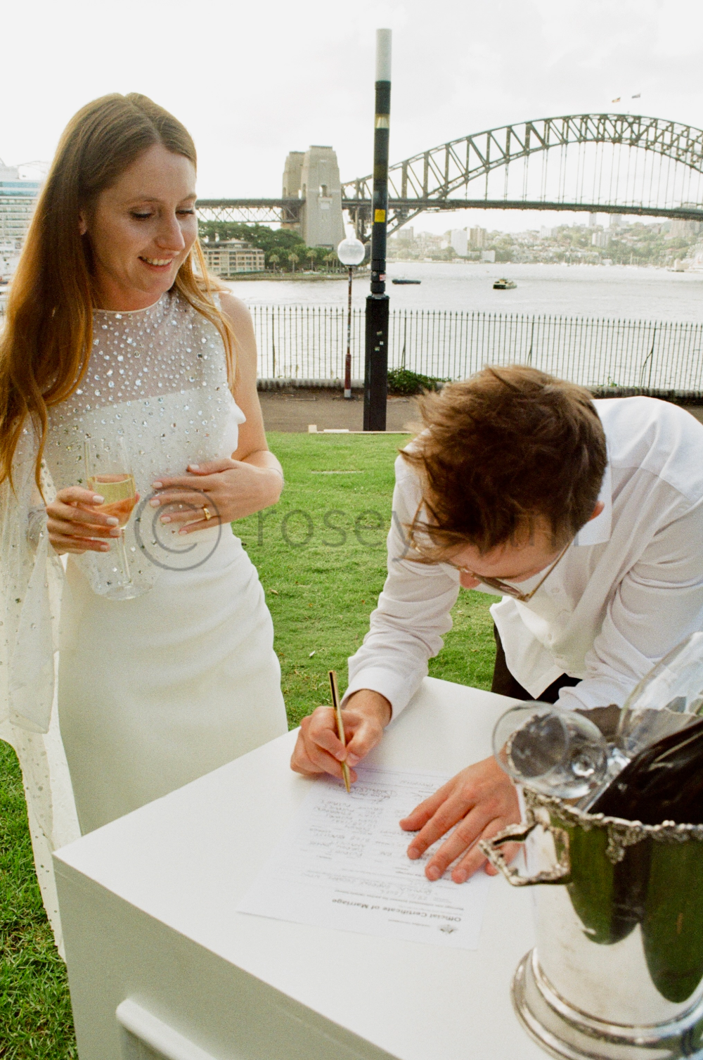 A woman in a white dress holding a glass of champagne smiling at a man signing a document on a table outdoors with the Sydney Harbour Bridge in the background.