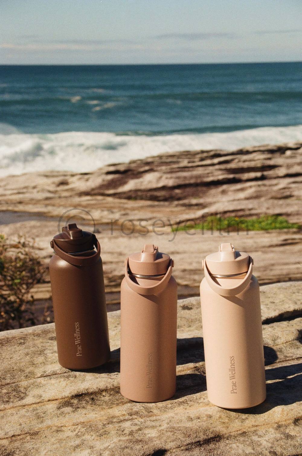 Three reusable water bottles in different shades of beige and brown on a rocky surface at the beach, with ocean waves and a cloudy sky in the background.