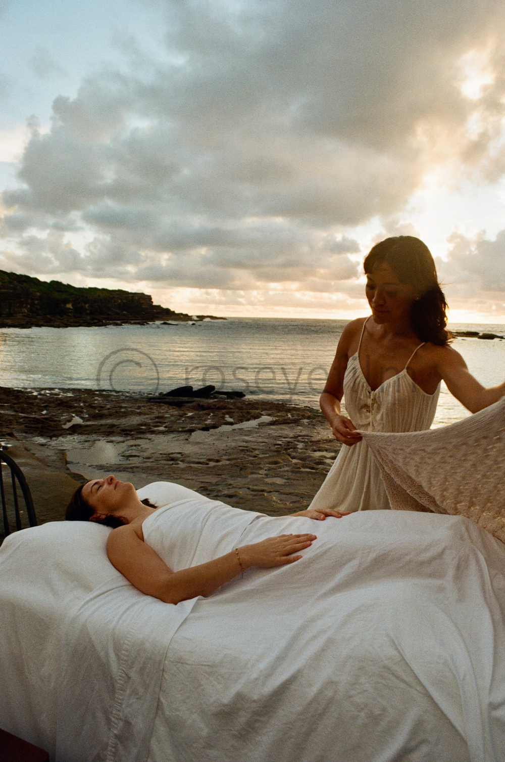 Two women on a beach at sunset, one lying on a bed and the other standing nearby holding a blanket.