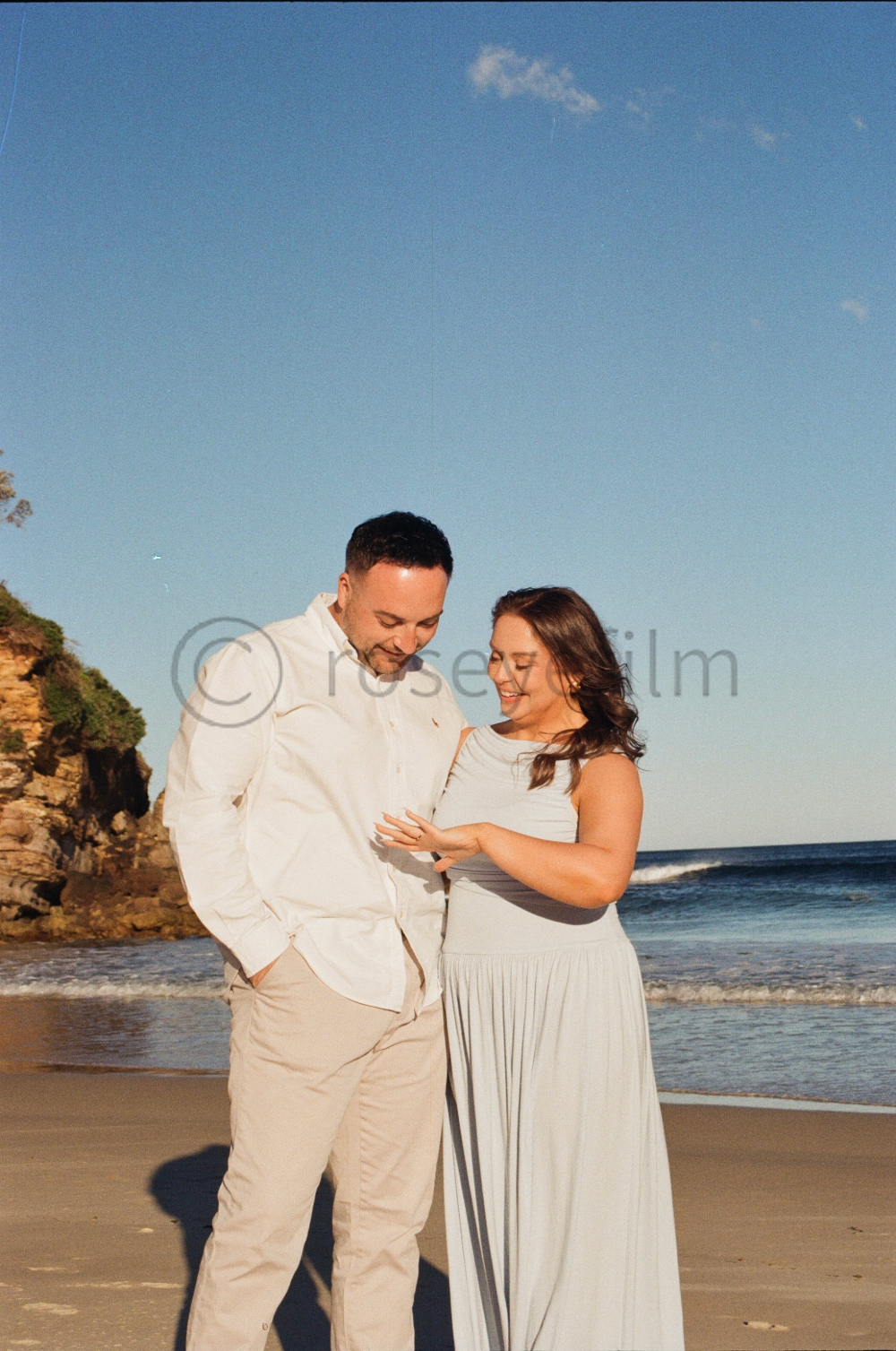 A smiling couple stands close together on a beach at sunset, with the ocean and a rocky cliff in the background.