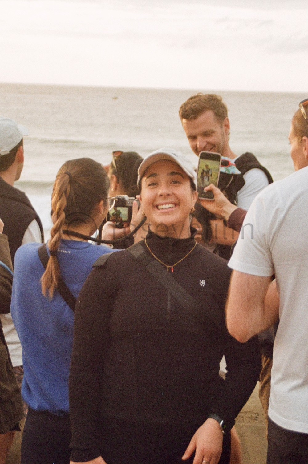 A group of people taking photos of a smiling woman on the beach, with ocean waves in the background.