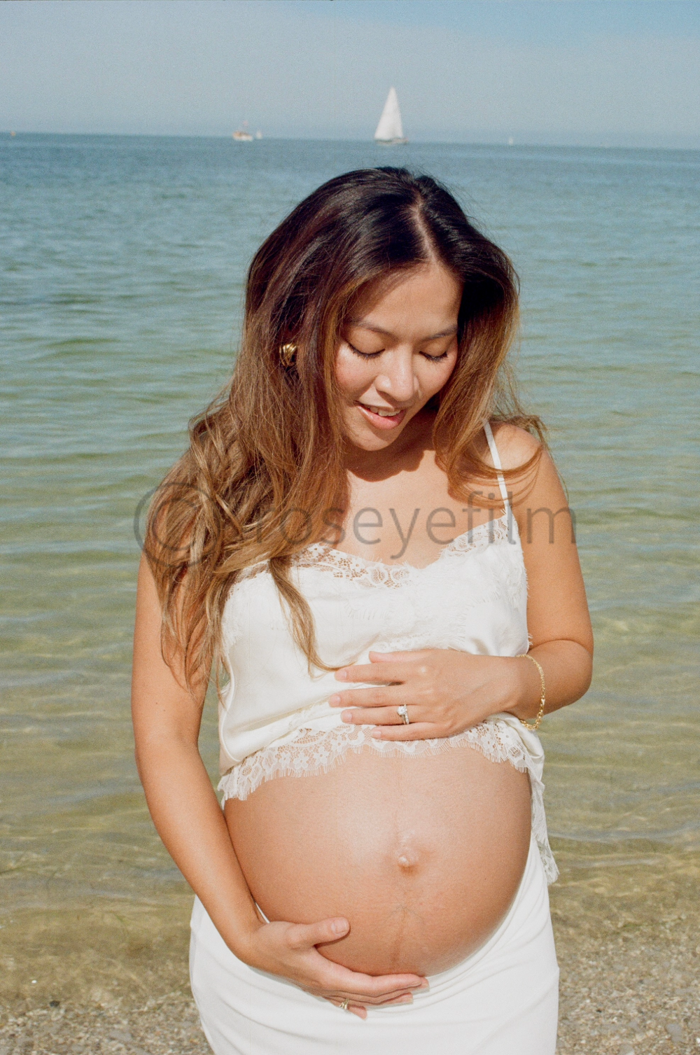 A pregnant woman standing at the beach, wearing a white lace top and skirt, touching her belly, with water and sailboats in the background.