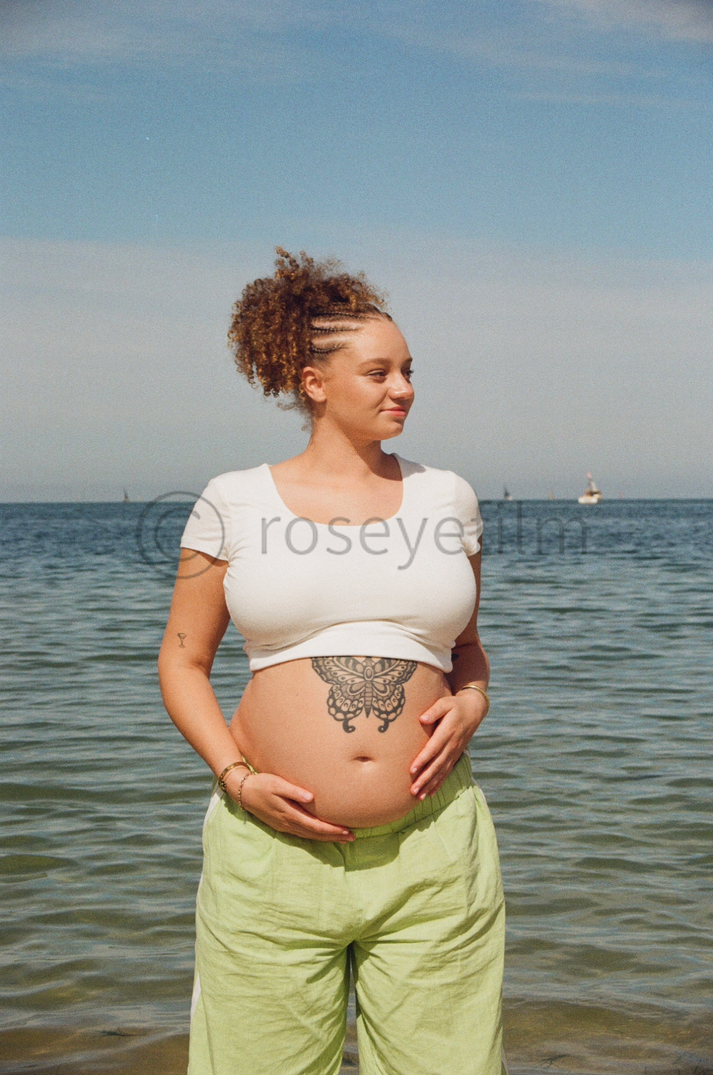 A pregnant woman with tattoos on her stomach and arms standing at the beach, holding her belly with both hands, wearing a white crop top and light green pants.