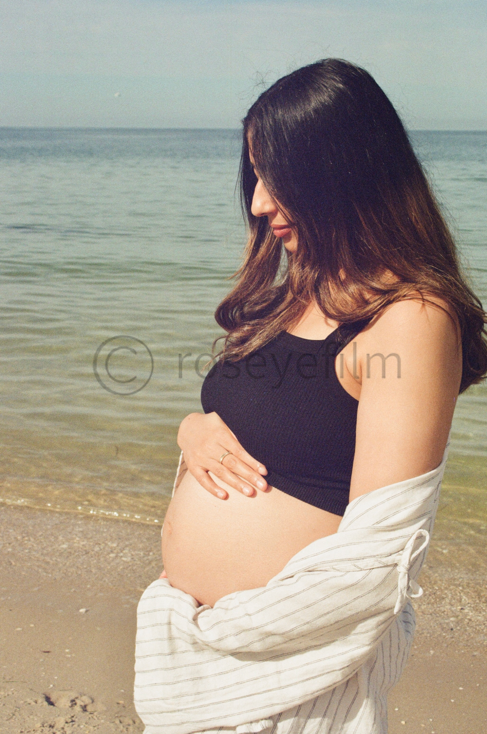 A pregnant woman stands on the beach near the water, gently resting her hand on her belly and looking down in profile, wearing a black top and a light-colored, striped jacket.