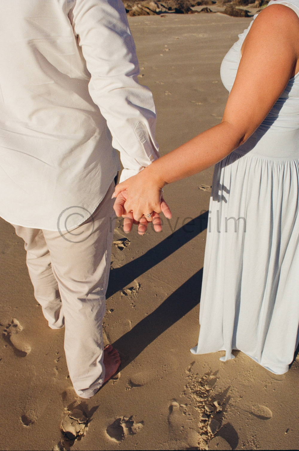 Close-up of a couple holding hands on a sandy beach, with the woman's hand featuring a ring and both dressed in white, suggesting a wedding or romantic occasion.