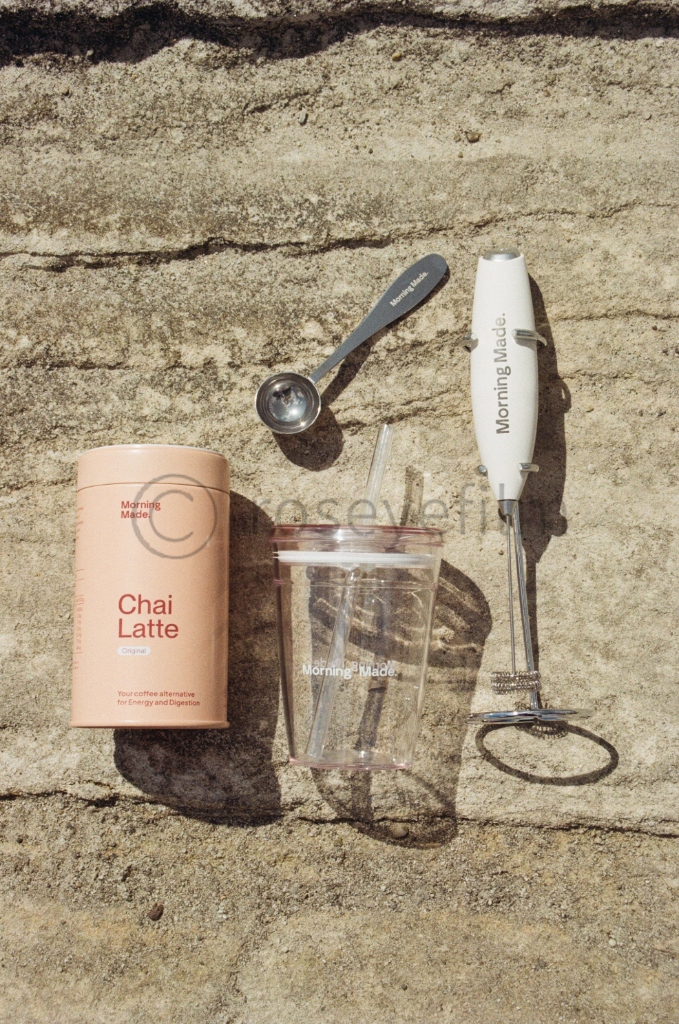 A container of Morning Made Chai Latte, a clear plastic cup with a straw, a small metal measuring spoon, and a hand-held milk frother, all placed on a sandy surface.