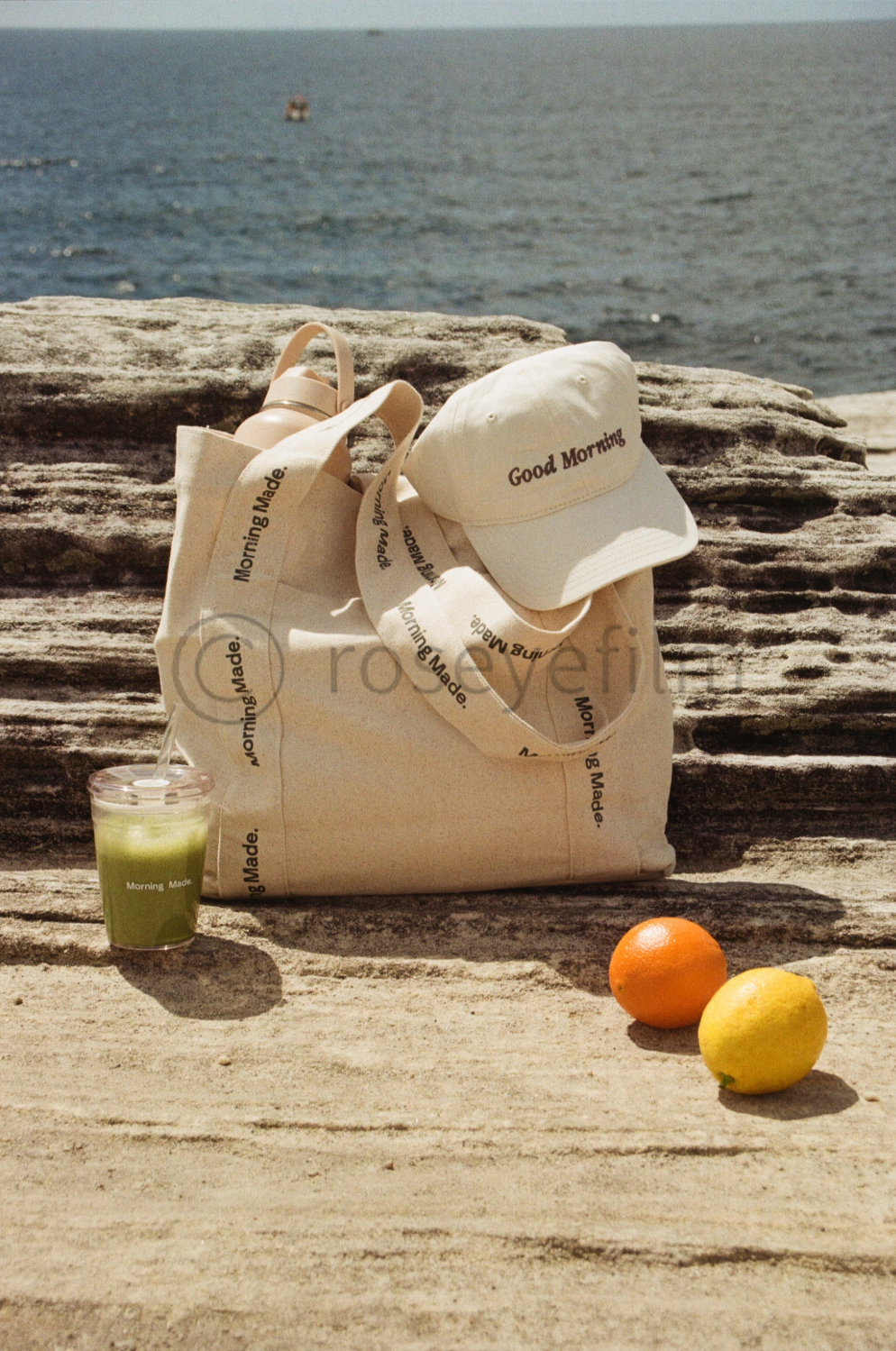 A beige tote bag with black text, a white cap with black text, a beige pair of sandals, a green beverage in a clear cup with a straw, an orange, and a lemon placed on a wooden surface near a body of water with a rocky shoreline in the background.