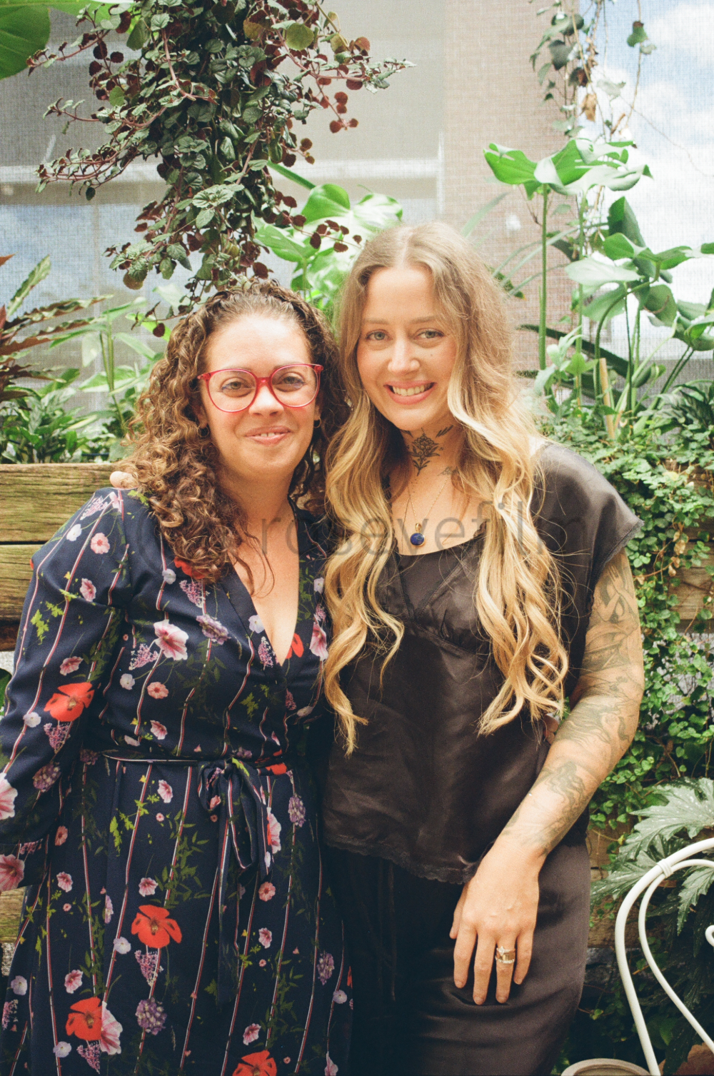 Two women standing close together in a lush, green garden. One is wearing a floral dress with red glasses, and the other is wearing a black top with tattoos and long blonde hair.