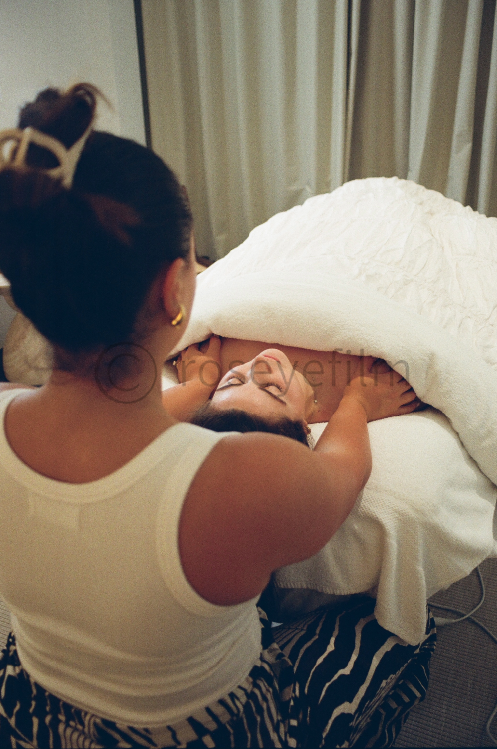 A woman giving a massage to another woman lying on a bed, covered with a white towel, in a room with beige curtains.