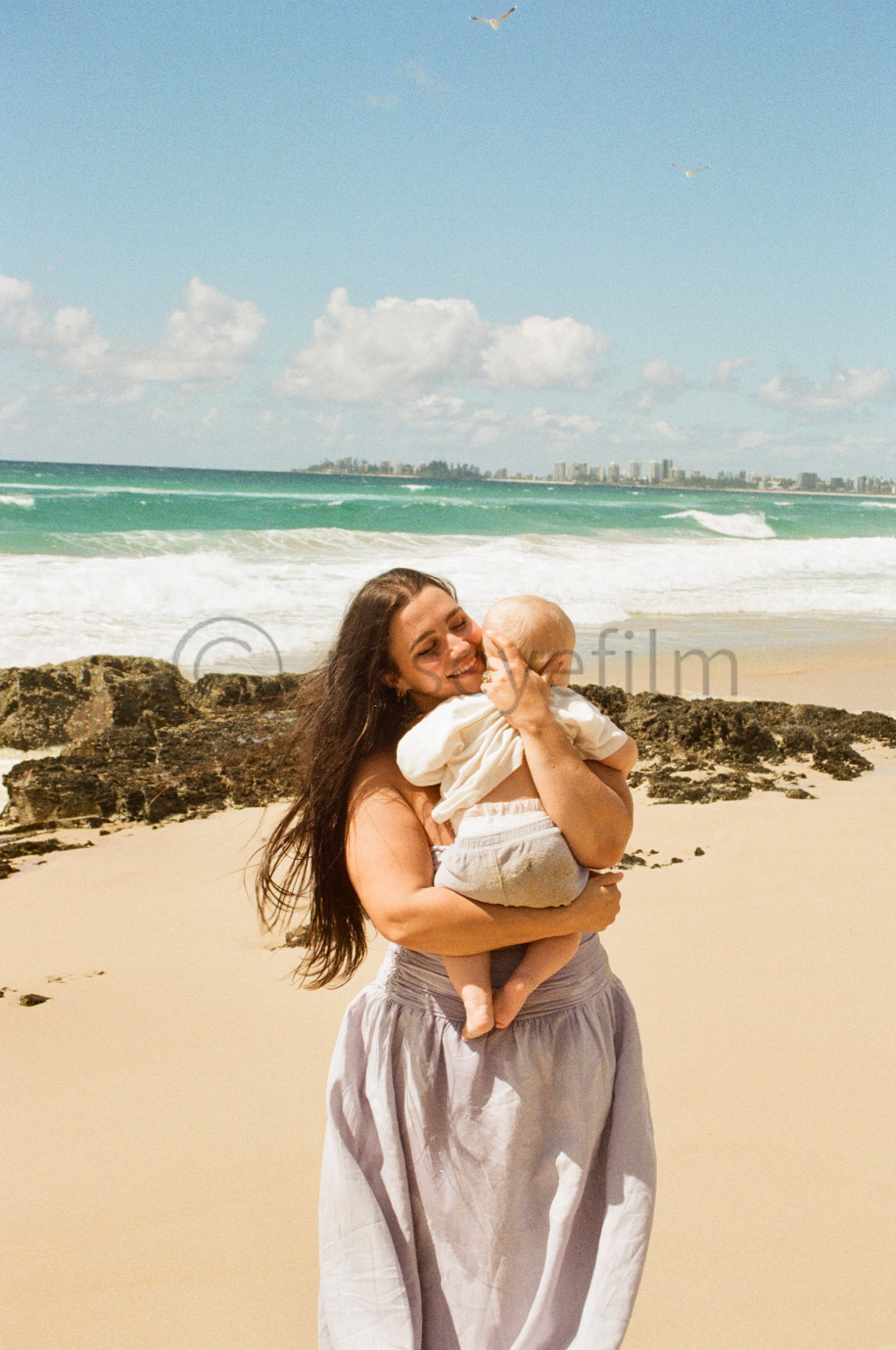 A woman with long dark hair holding a small child on a sandy beach with rocks, ocean waves, and a cityscape in the background, under a partly cloudy sky.