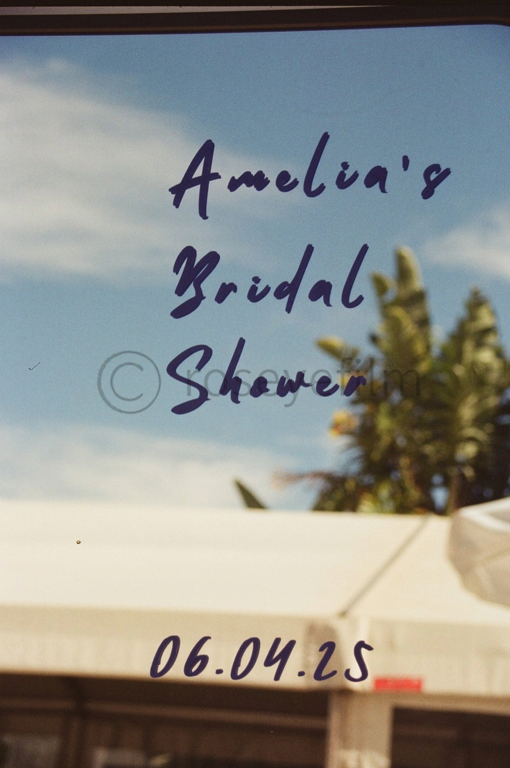 Sign on glass door reading 'Amelia's Bridal Shower' with date '06.04.25' underneath, outside sky with clouds and trees visible.