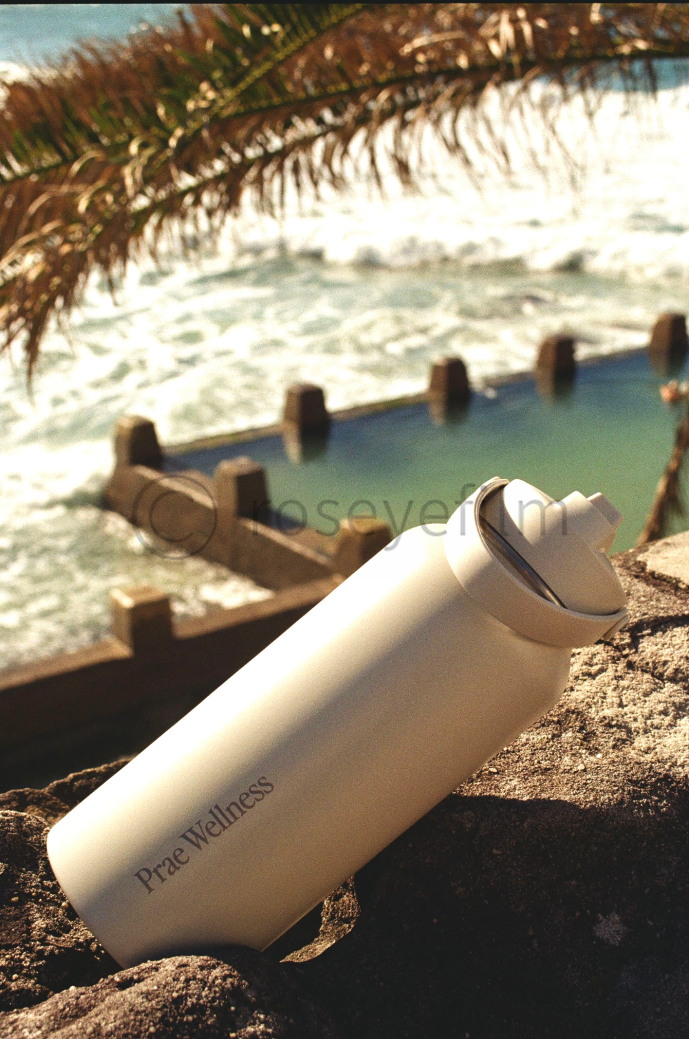 A white travel mug with 'Pra Wellness' written on it, resting on a rock with a beach and waves in the background.