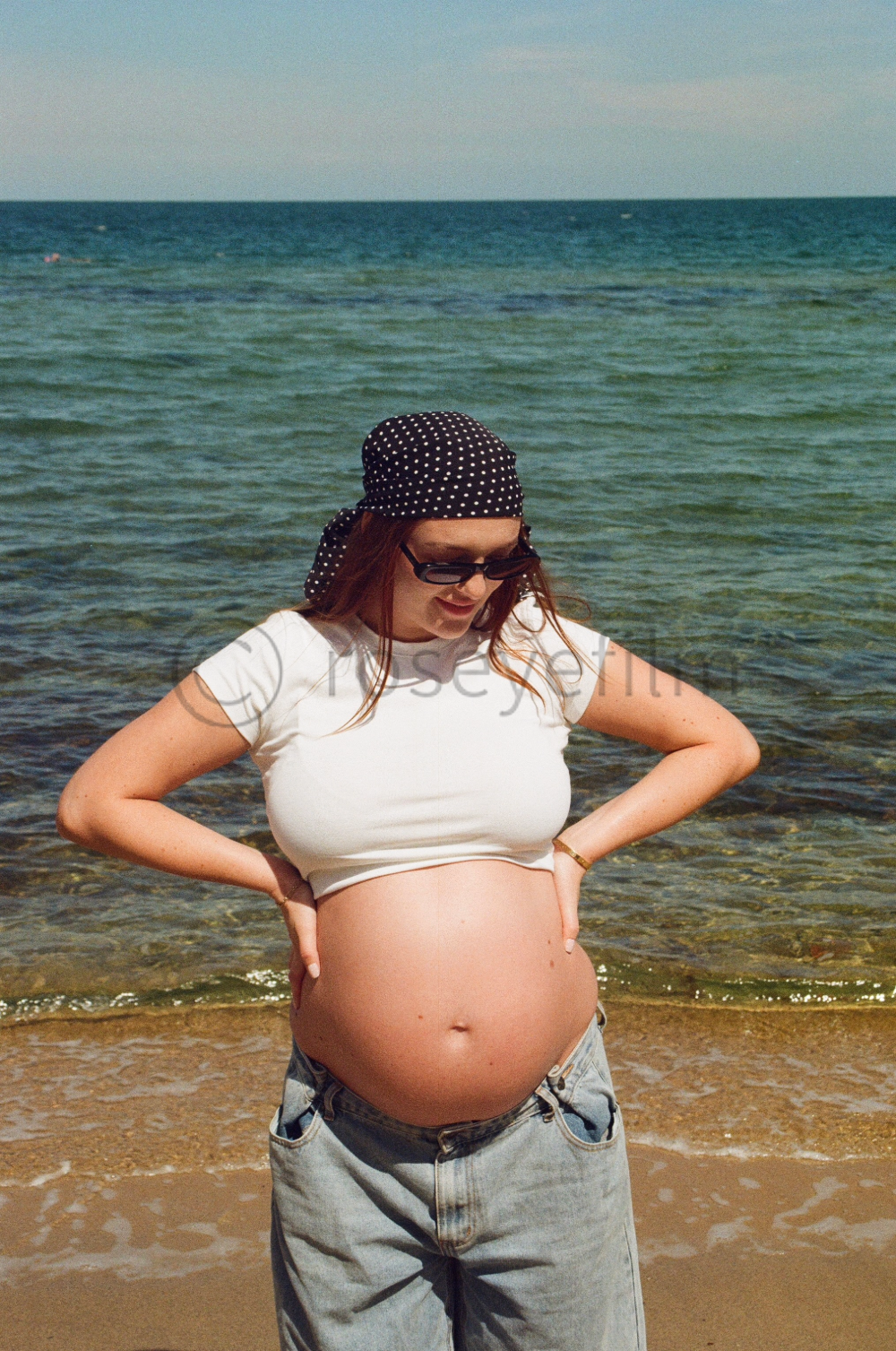 Pregnant woman standing on the beach with hands on her hips, wearing sunglasses, a white t-shirt, and a black headscarf with white polka dots, looking down at her belly.