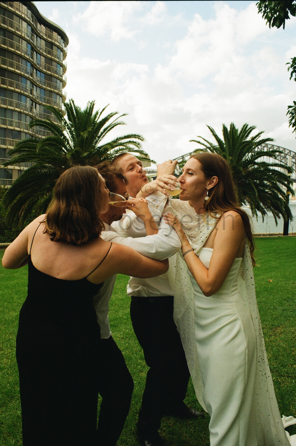 A group of four people, two women and two men, including a bride and groom, celebrating outdoors on a grassy area with palm trees and a cityscape in the background. They are laughing and sharing drinks.