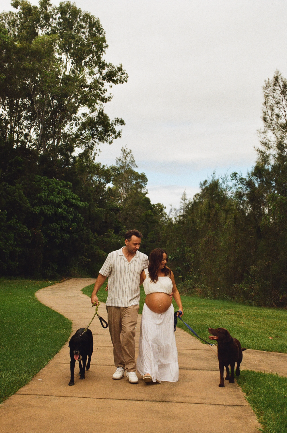 A pregnant woman and her partner smiling and walking on a park path with two dogs, surrounded by lush green trees during daytime.