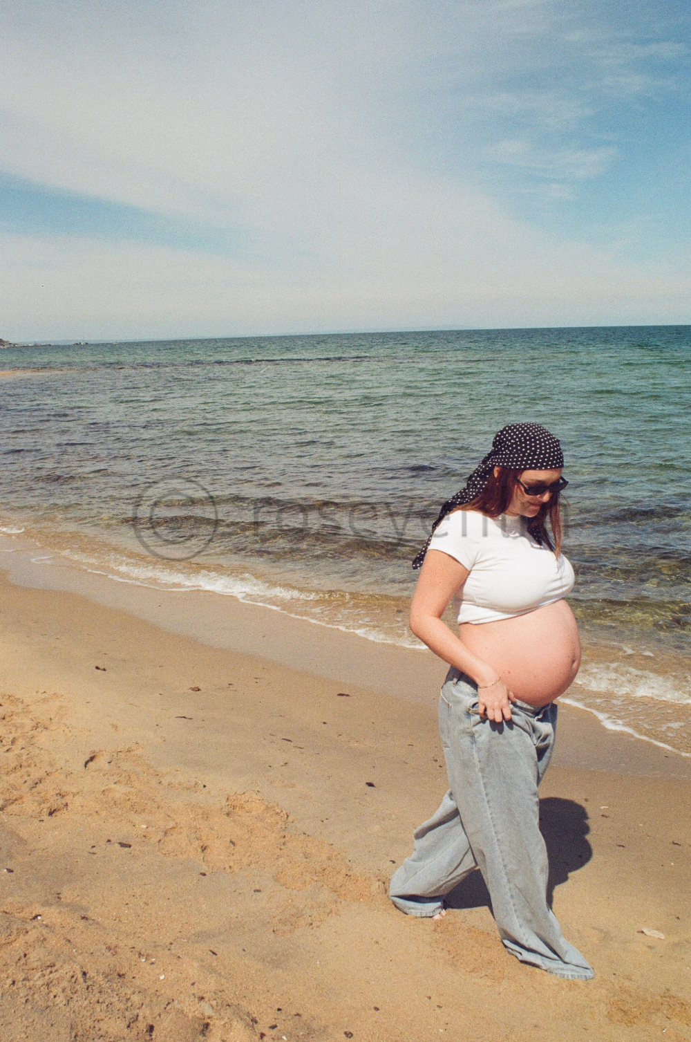 A pregnant woman in a white t-shirt, loose jeans, sunglasses, and a black polka dot headscarf walking on a sandy beach near the ocean during daytime.