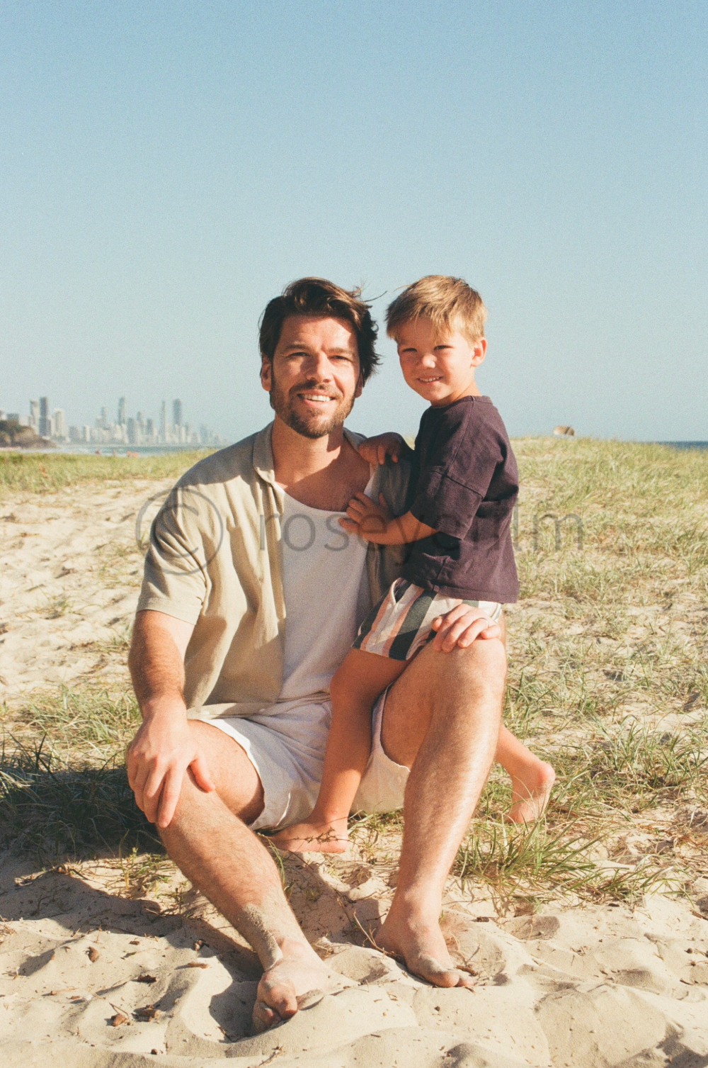 A man and a young boy sitting on the sandy beach near grass, with the city skyline in the background, enjoying a sunny day.