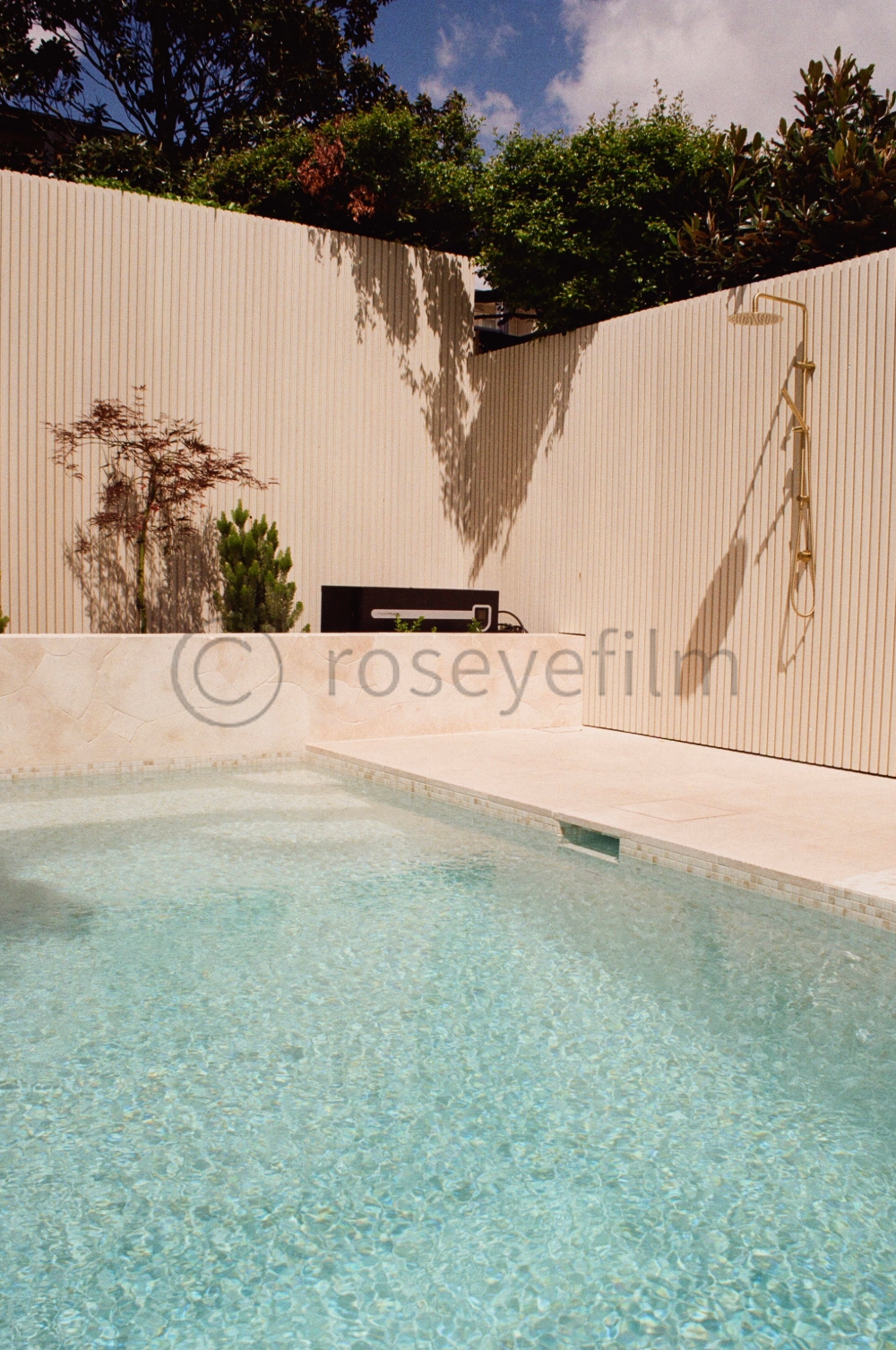 An outdoor swimming pool with clear water, surrounded by a beige patio area. There is a beige privacy wall with shadows of trees cast on it, and a garden with small trees and plants. An outdoor shower is mounted on the wall.