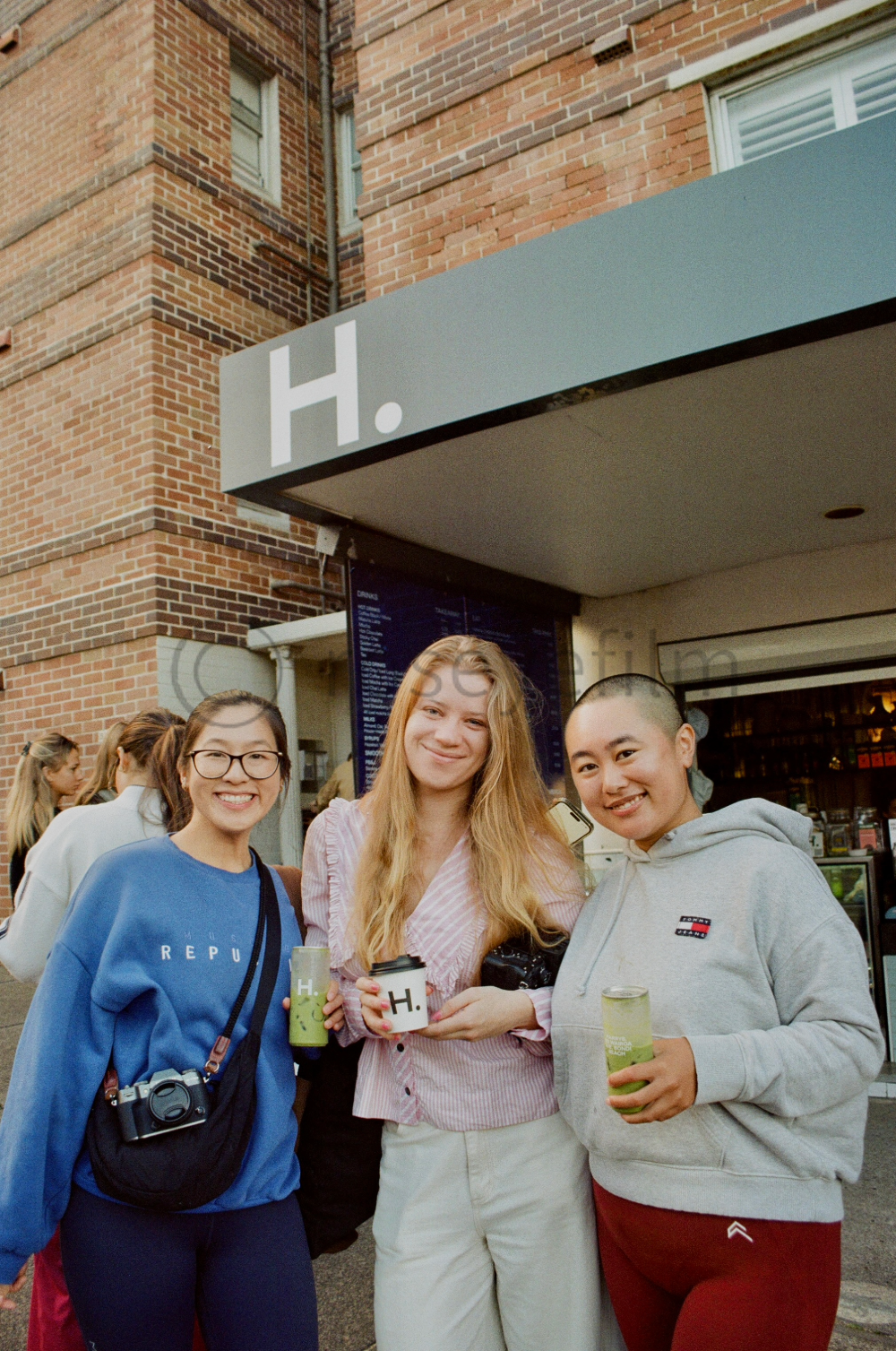 Three young women standing outside a cafe, holding drinks, smiling at the camera. One woman wears glasses and a blue sweatshirt, another has long blonde hair and a pink striped shirt, and the third has a buzz cut and a gray hoodie. The cafe has a mod