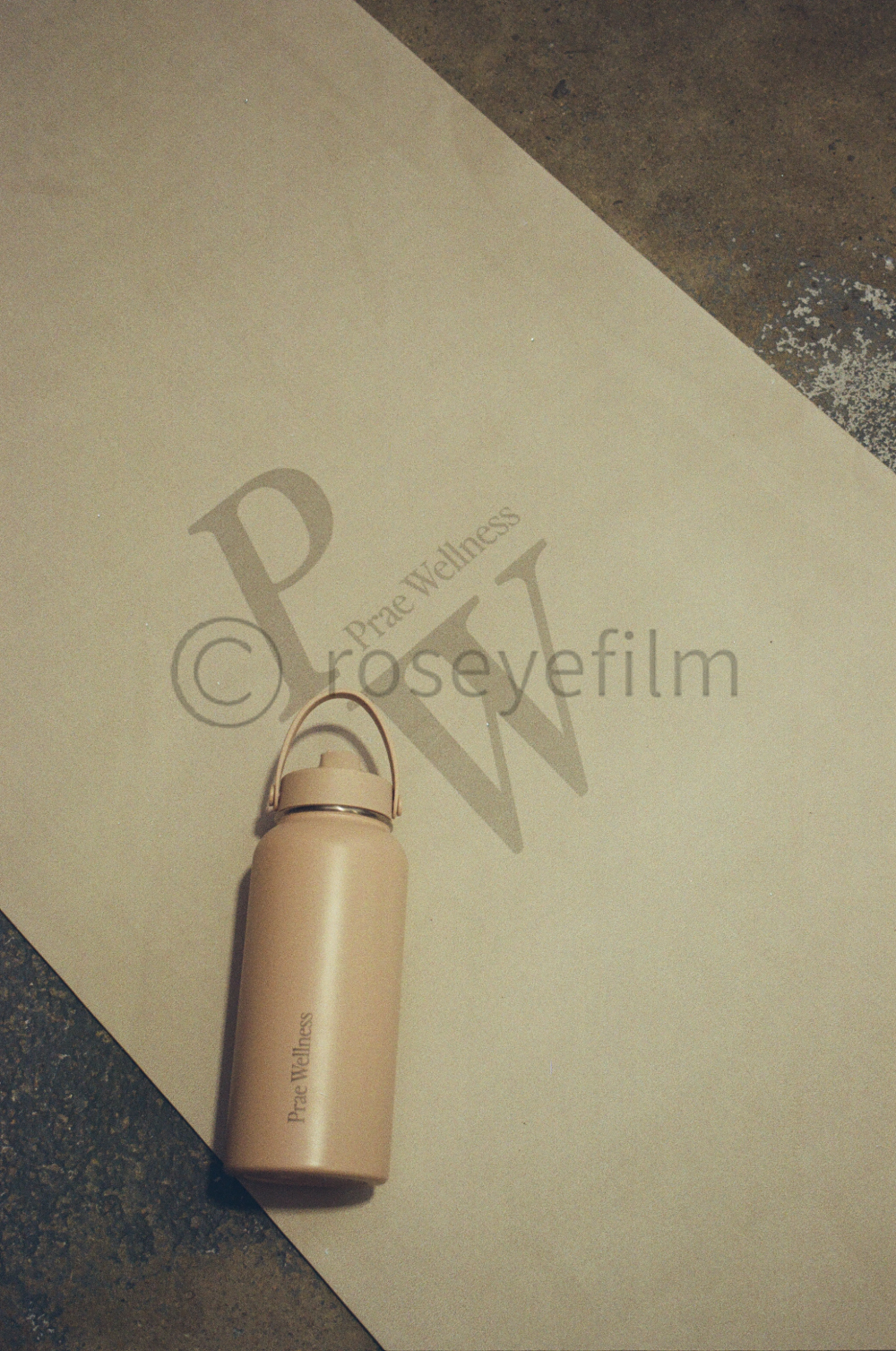 A beige water bottle with a handle on top, labeled 'Praec Wellness,' placed on a light-colored surface that has a shadow with the words 'Praec Wellness' and 'L' and 'W' visible, with a darker textured floor at the bottom and top right corner.