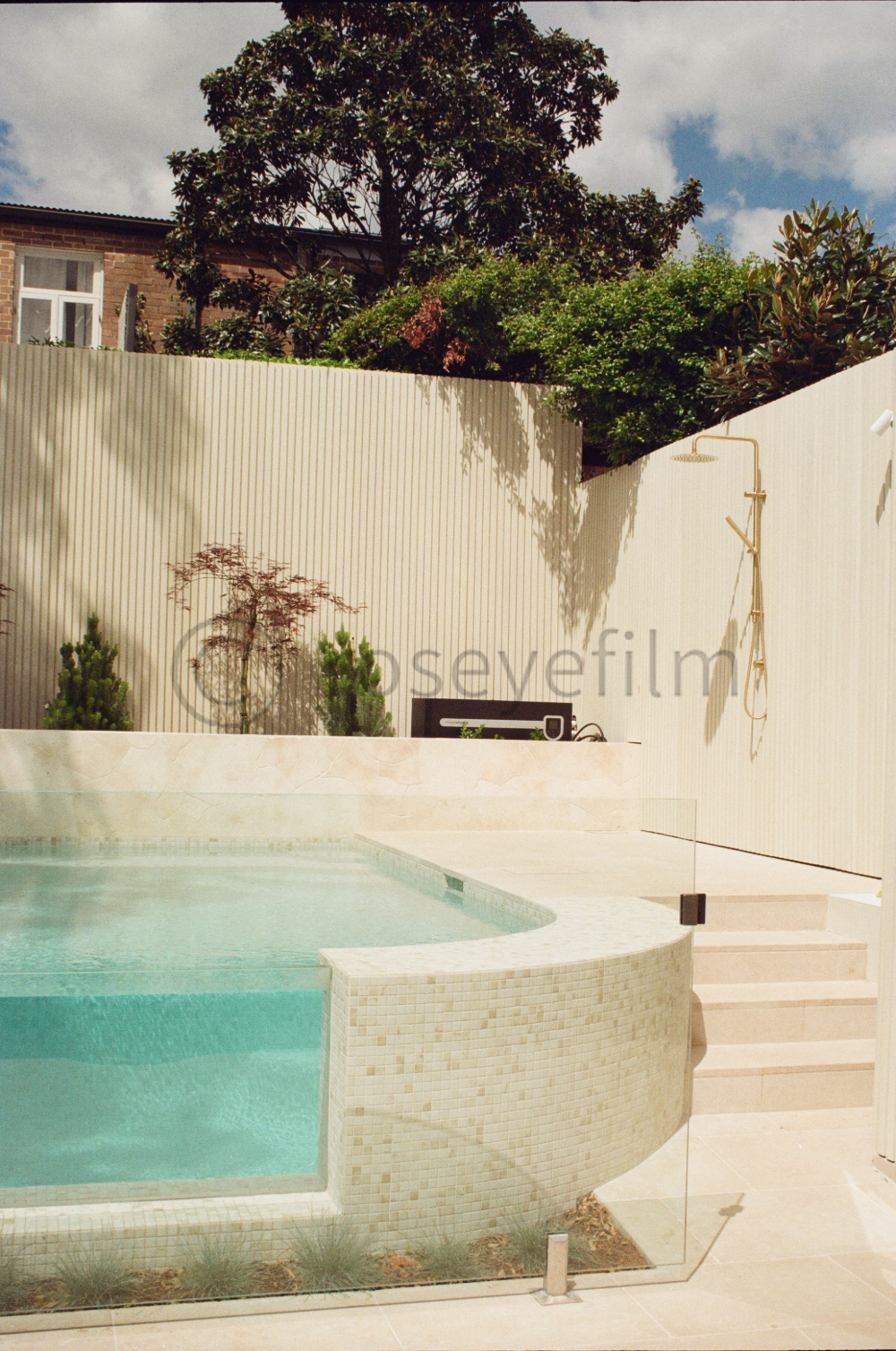 A modern outdoor swimming pool with light-colored tile surrounded by a privacy wall, small plants, and a staircase leading to a higher level. There is an outdoor shower mounted on the wall and a black timer device on the ledge above the pool. The bac