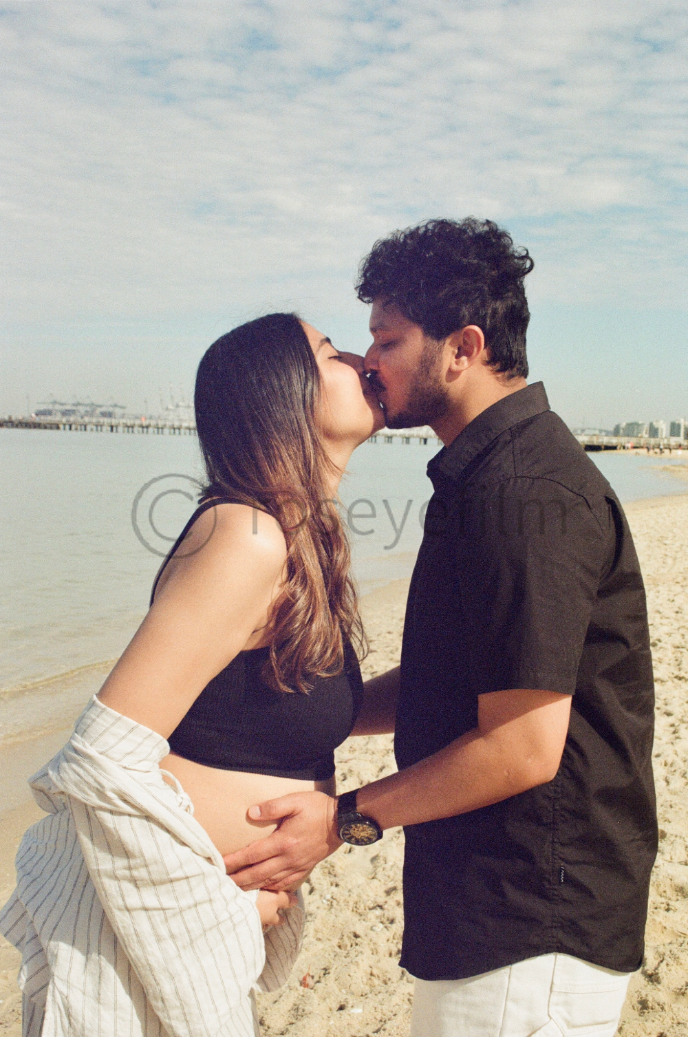 A couple kissing on the beach near the water, with a pier and city skyline in the background.