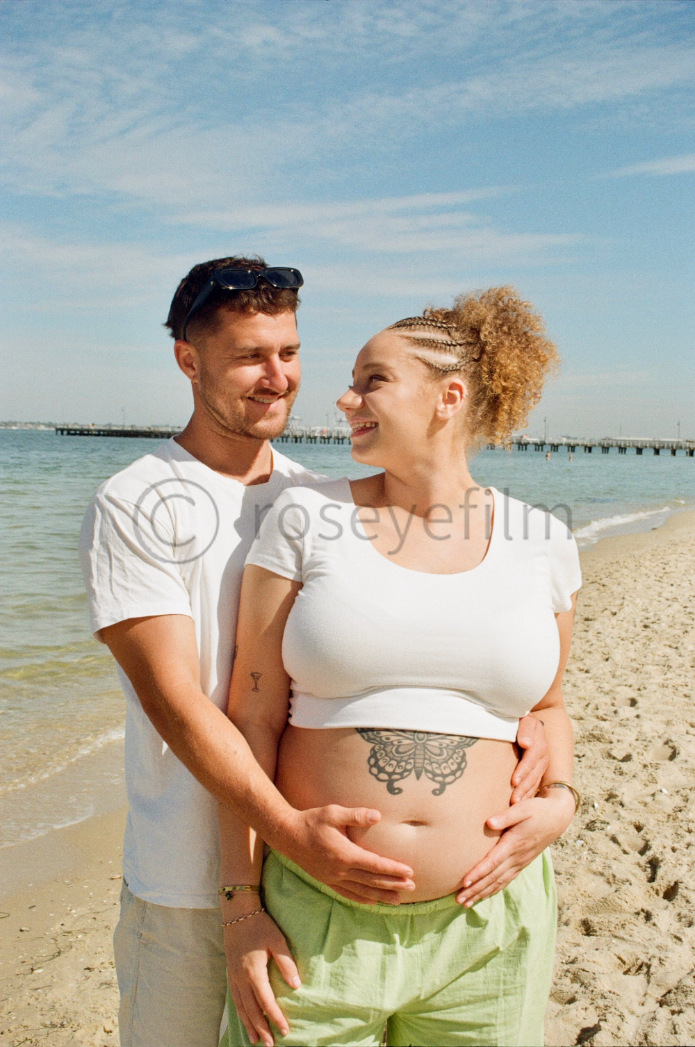A pregnant woman and her partner standing on a beach, with the woman showing her tattooed belly and smiling back at her partner. The man is standing behind her, gently holding her belly, both are smiling and enjoying a sunny day by the water.