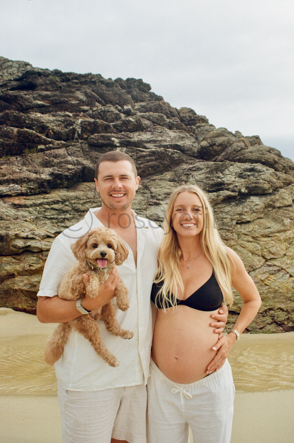 A happy couple stands on a beach with rocky cliffs behind them. The pregnant woman wears a black bikini top and white pants, while the man in a white shirt and trousers holds a small, curly-haired dog. The woman has long blonde hair, and the man has 