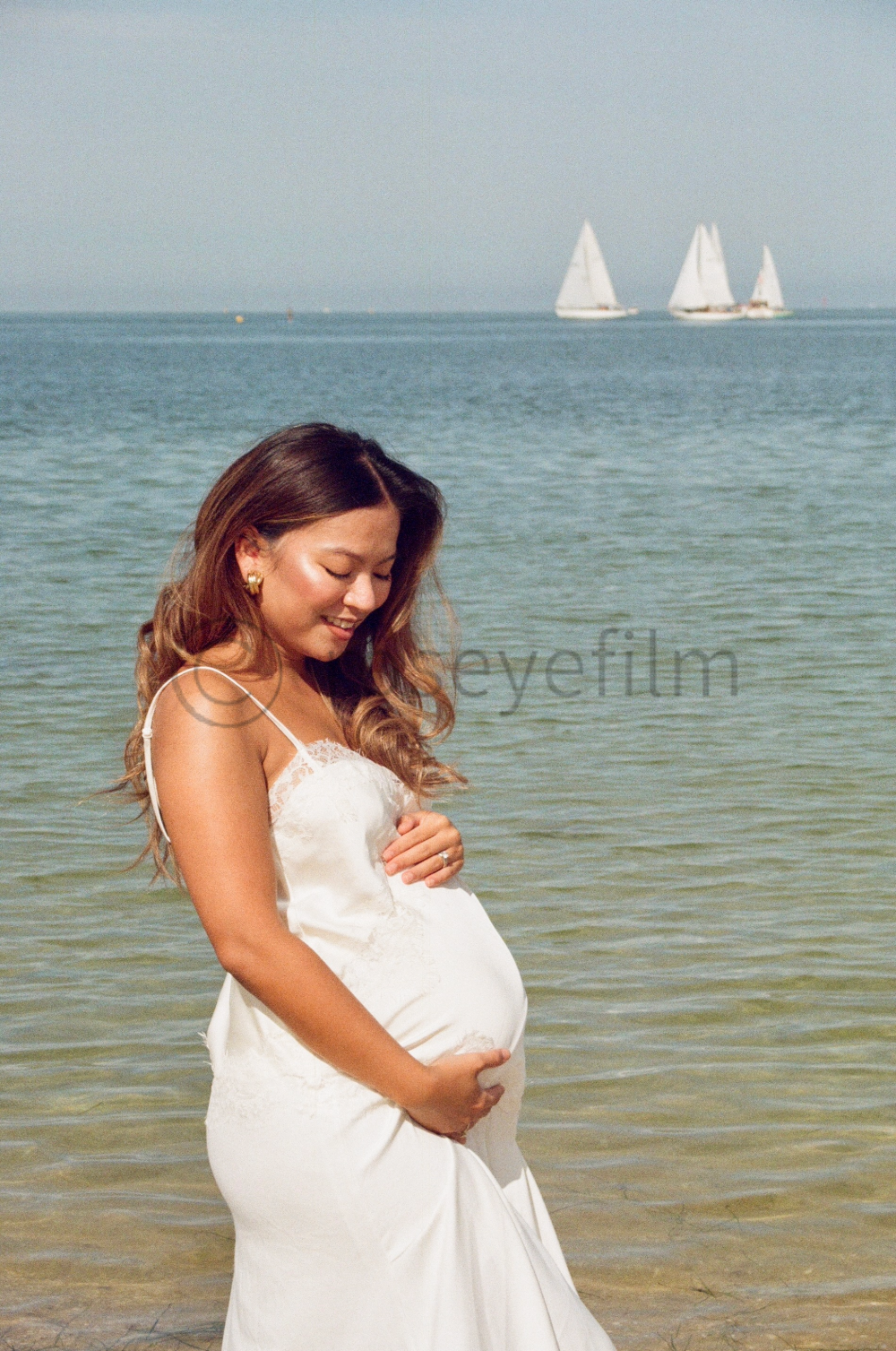 Pregnant woman in a white dress standing in shallow ocean water with sailboats in the background.