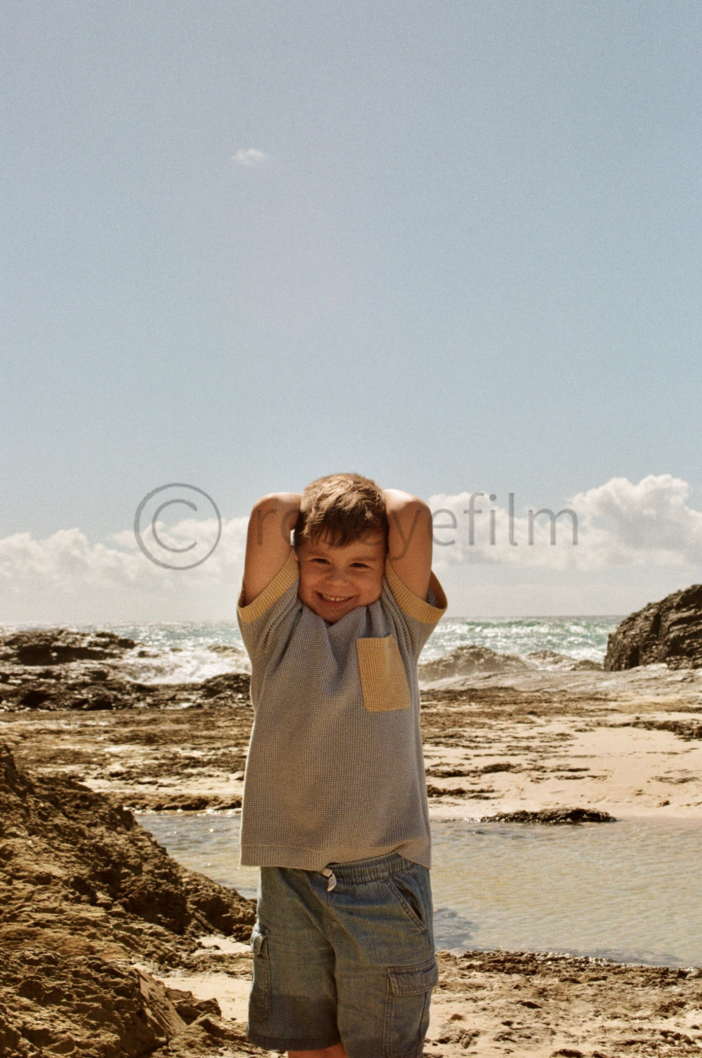 A young boy smiling with hands behind his head on a beach with rocks, sand, and ocean waves in the background under a partly cloudy sky.