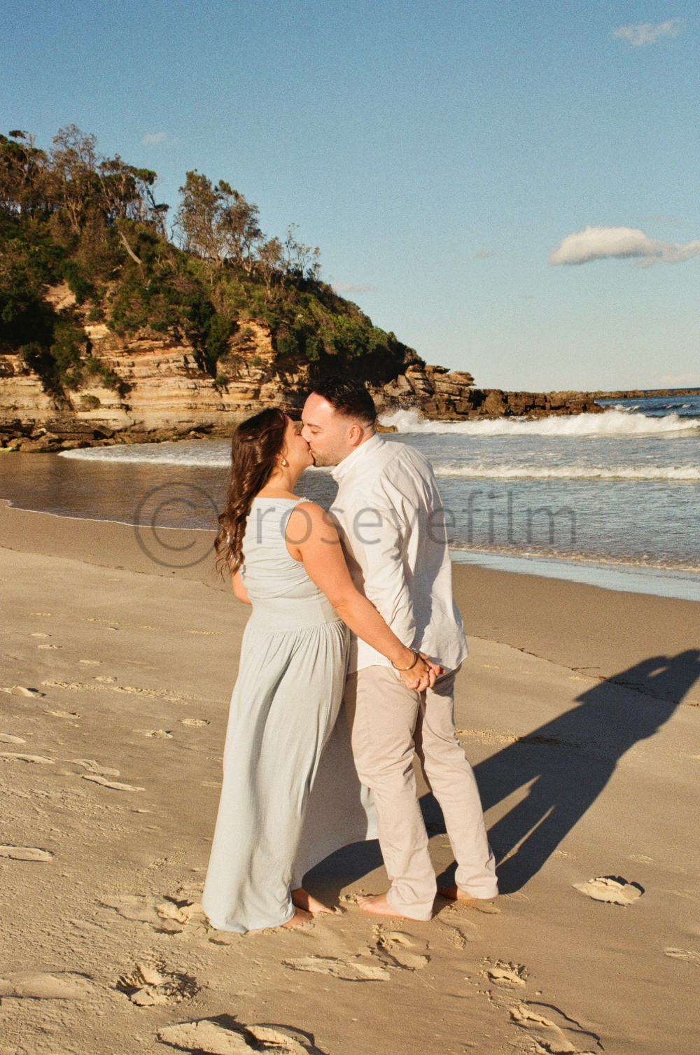 A couple kissing on a sandy beach with cliffs in the background and ocean waves.