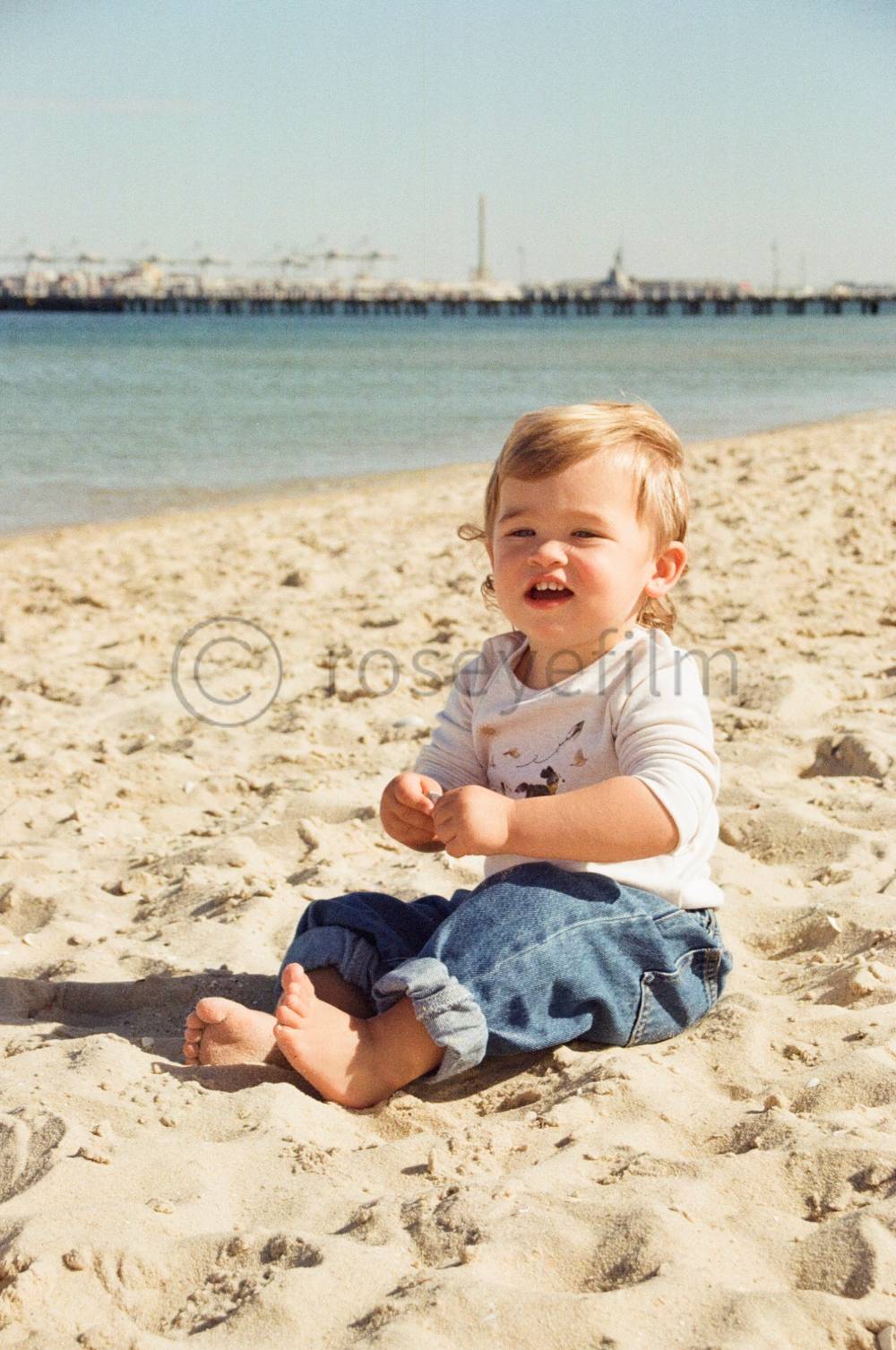 A young child sitting on a sandy beach near the water with a pier in the background.