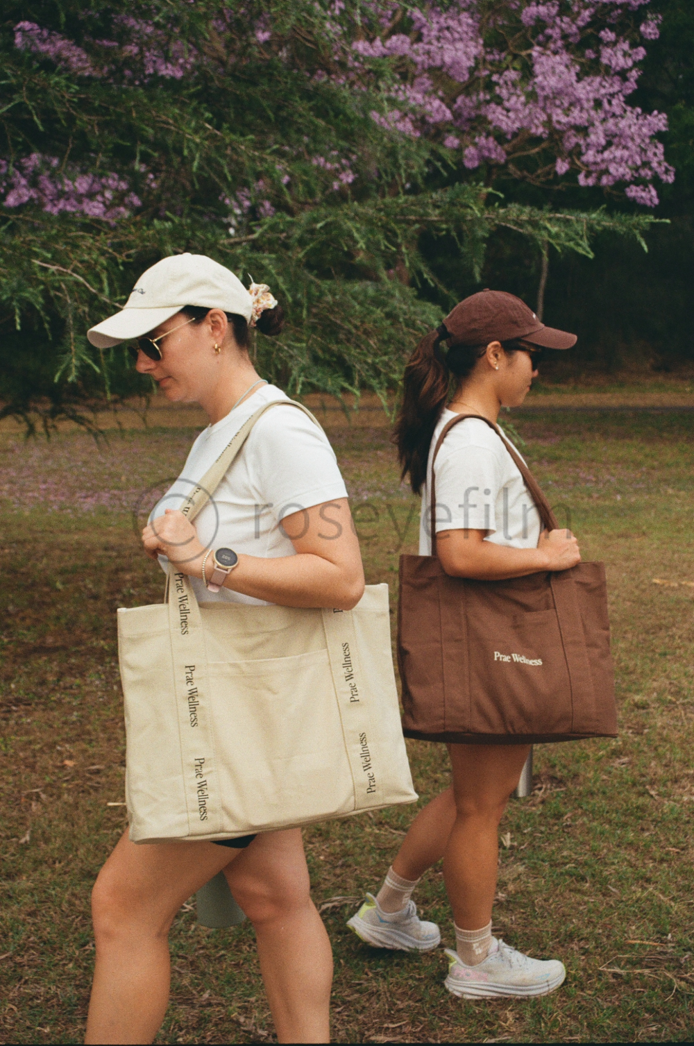 Two young women walking outdoors during daytime, carrying large tote bags with the words "Pree Wellness" on them. They are wearing casual sportswear, caps, and sunglasses, and are surrounded by greenery and blooming trees with purple flowers.