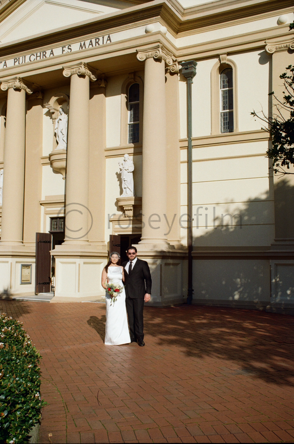 A bride and groom standing outside a church, dressed in wedding attire, with the bride holding a bouquet of flowers.