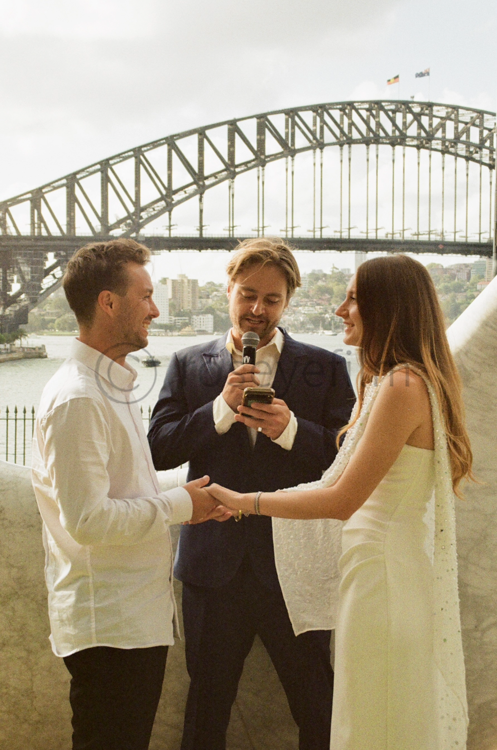 A wedding ceremony taking place outdoors with the Sydney Harbour Bridge in the background, featuring a man and woman exchanging vows while holding hands, and an officiant reading from a phone.
