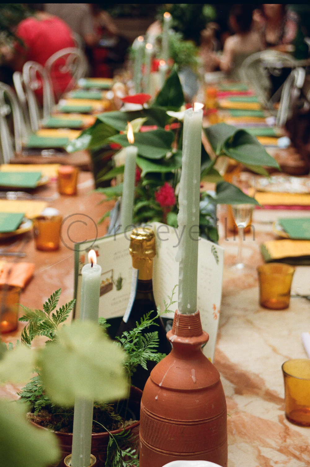 A decorated dining table set for a celebration with green candles, potted plants, amber glassware, and a terracotta bottle in the foreground. In the background, guests are sitting and chatting, with colorful table settings and blurred greenery.