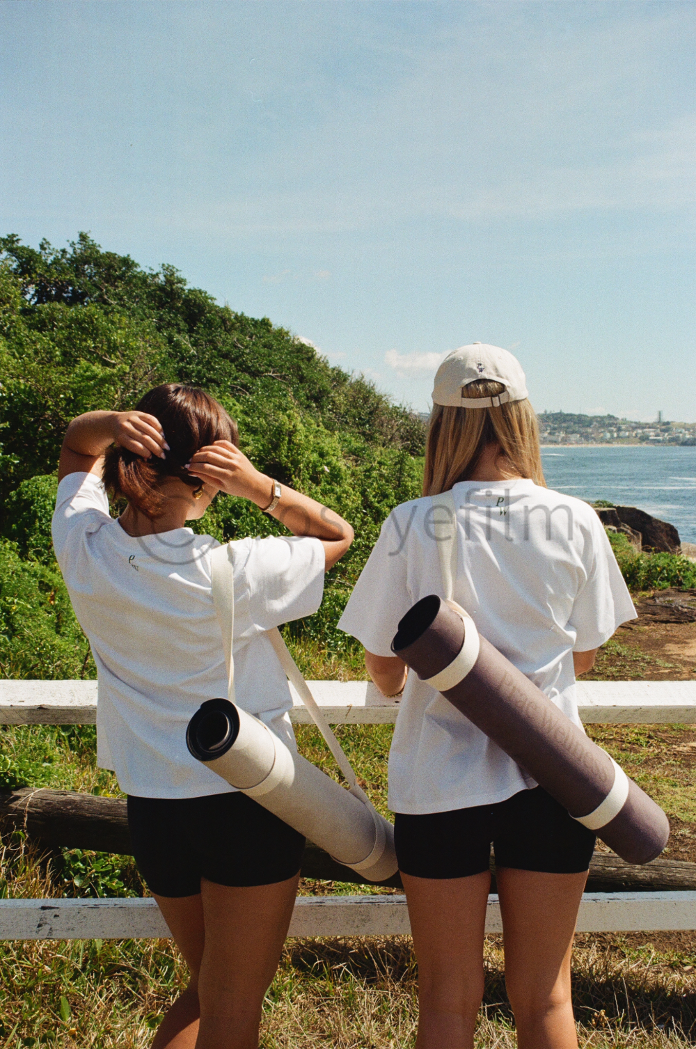 Two women with yoga mats on their backs standing outdoors by a white fence, overlooking greenery and water, with a cityscape in the distance under a blue sky.
