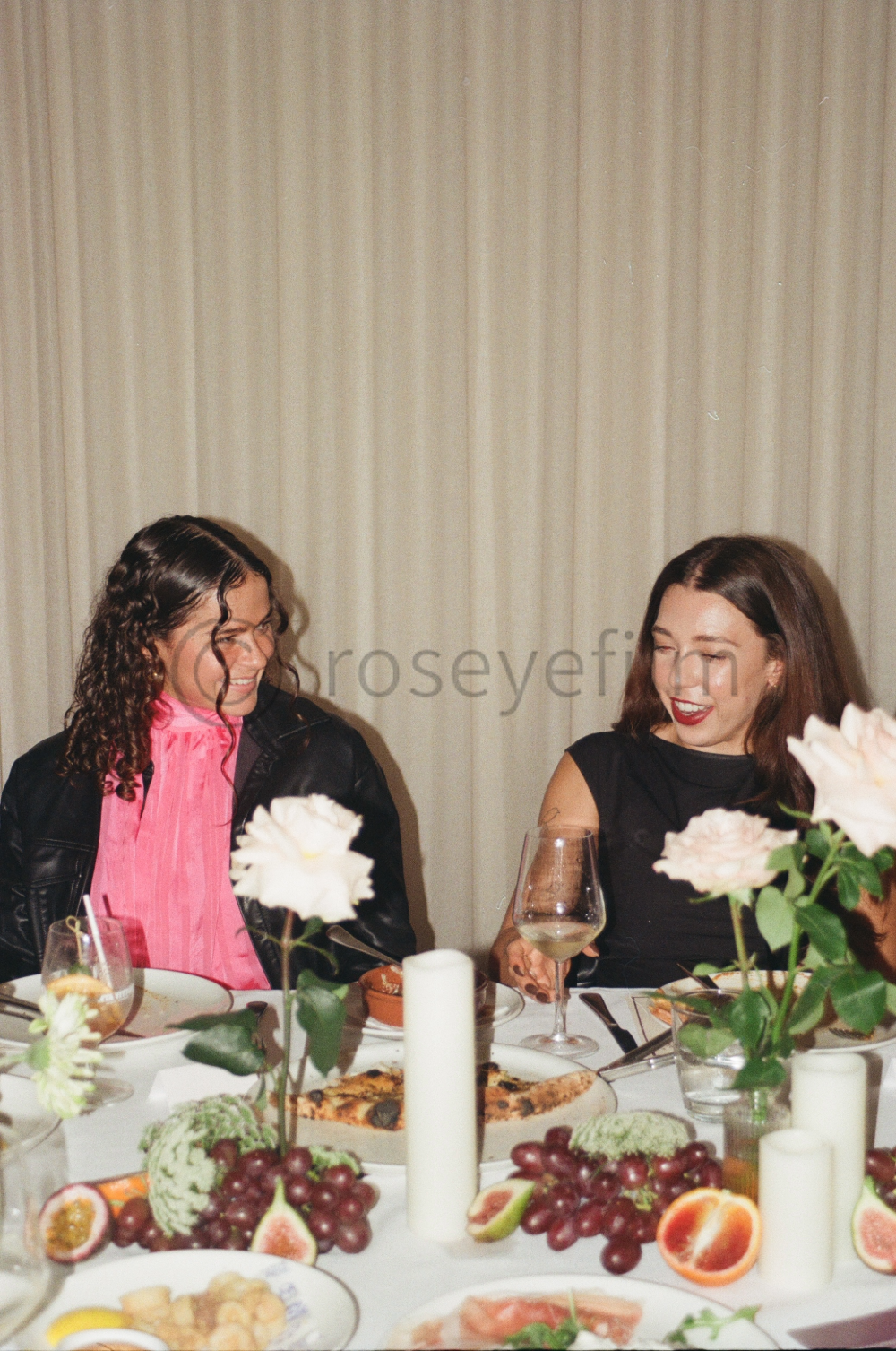 Two women smiling and talking at a dinner party with a table of food, flowers, candles, and glasses of wine.