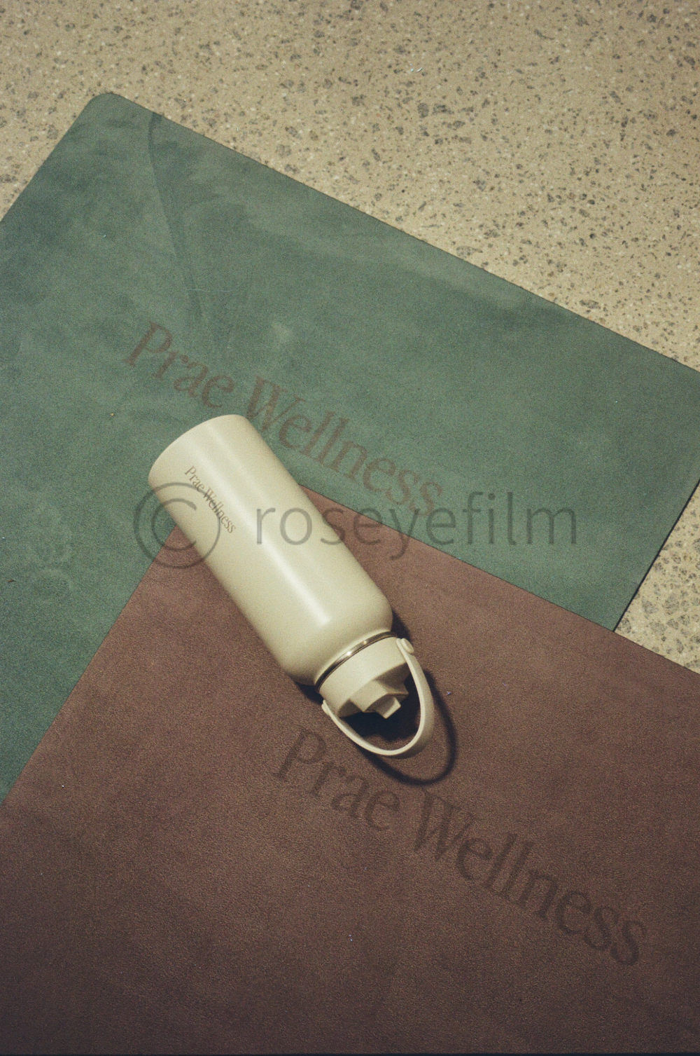 A white Prac Wellness water bottle resting on green and brown yoga mats labeled "Peace Wellness" on a speckled floor.