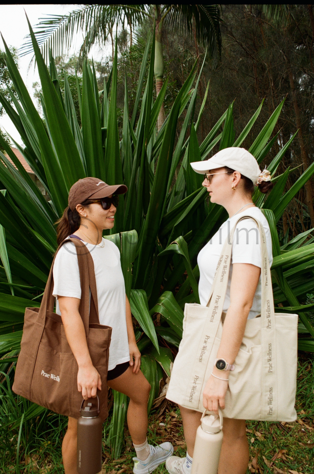 Two women standing outdoors near tall green plants, wearing casual sportswear, sunglasses, and caps, carrying tote bags and water bottles, talking and smiling.