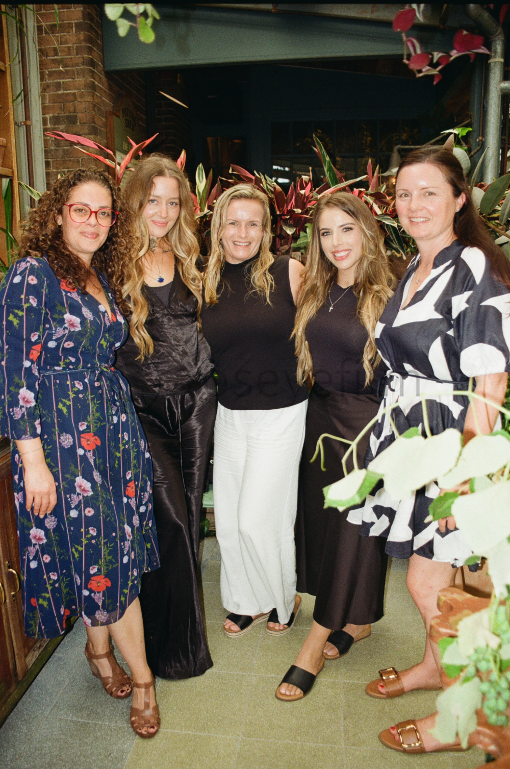 Five women standing together, smiling, in a floral-patterned dress, leather pants, black top with white pants, black skirt, and a black and white patterned dress, surrounded by indoor plants, in a cozy restaurant or cafe setting.