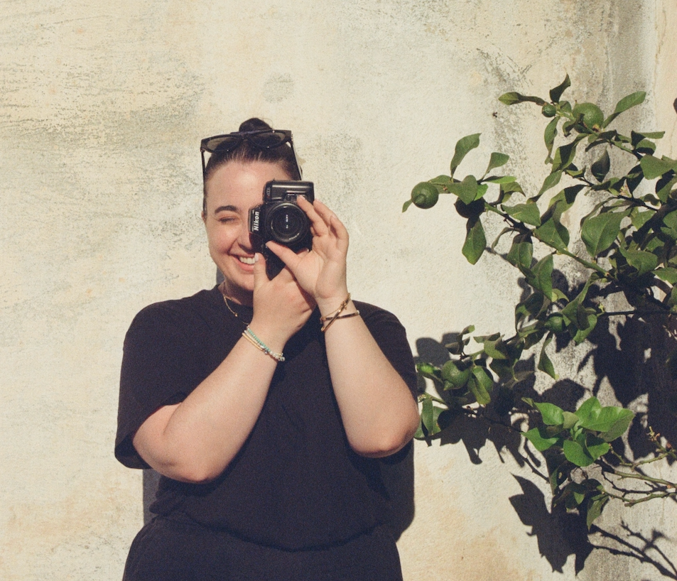 A young woman in a black shirt taking a selfie with a camera, smiling and standing next to a green leafy plant against a textured white wall.
