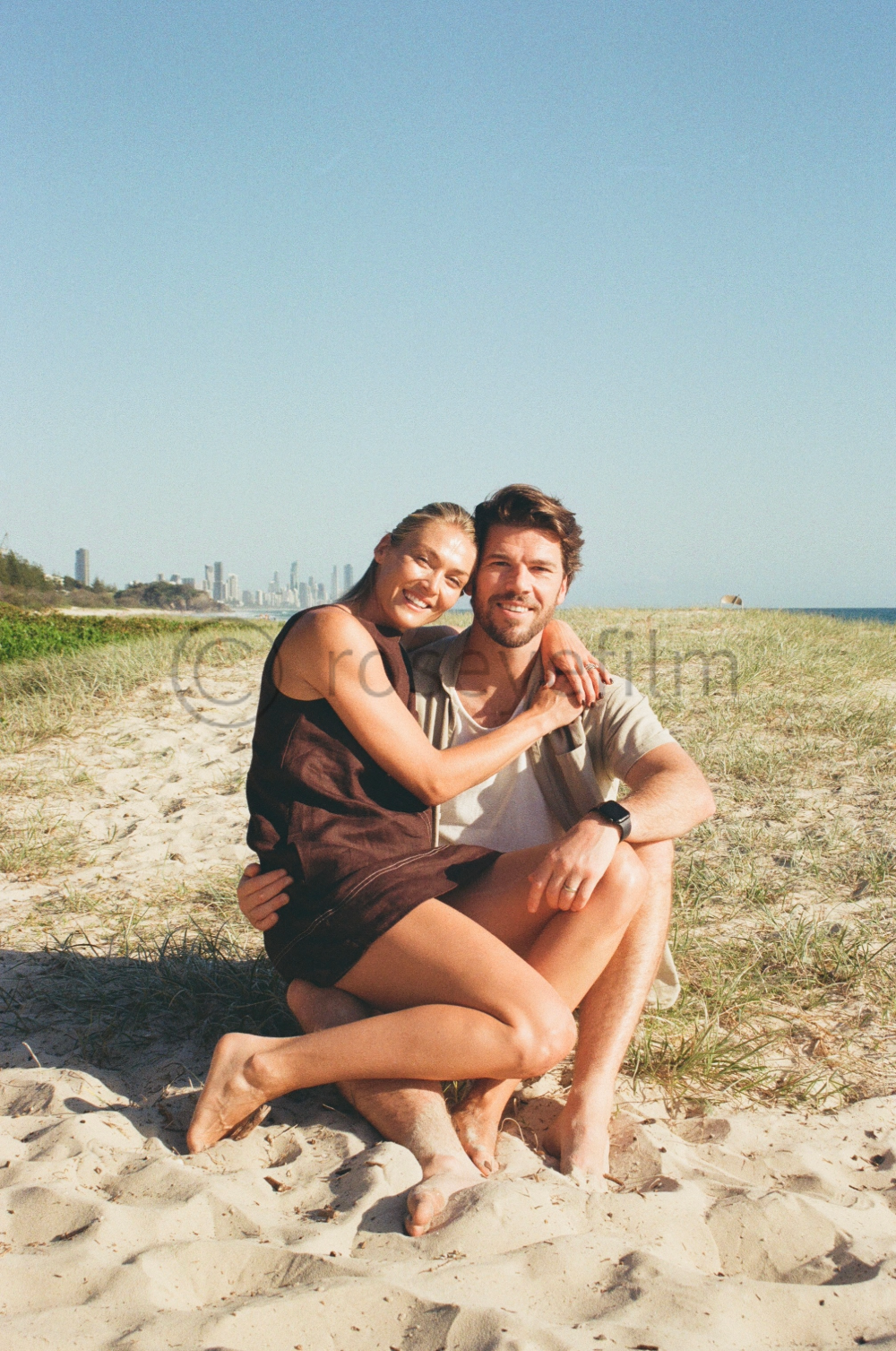 A couple sitting on the sand at the beach, smiling and embracing, with city skyline in the background on a sunny day.