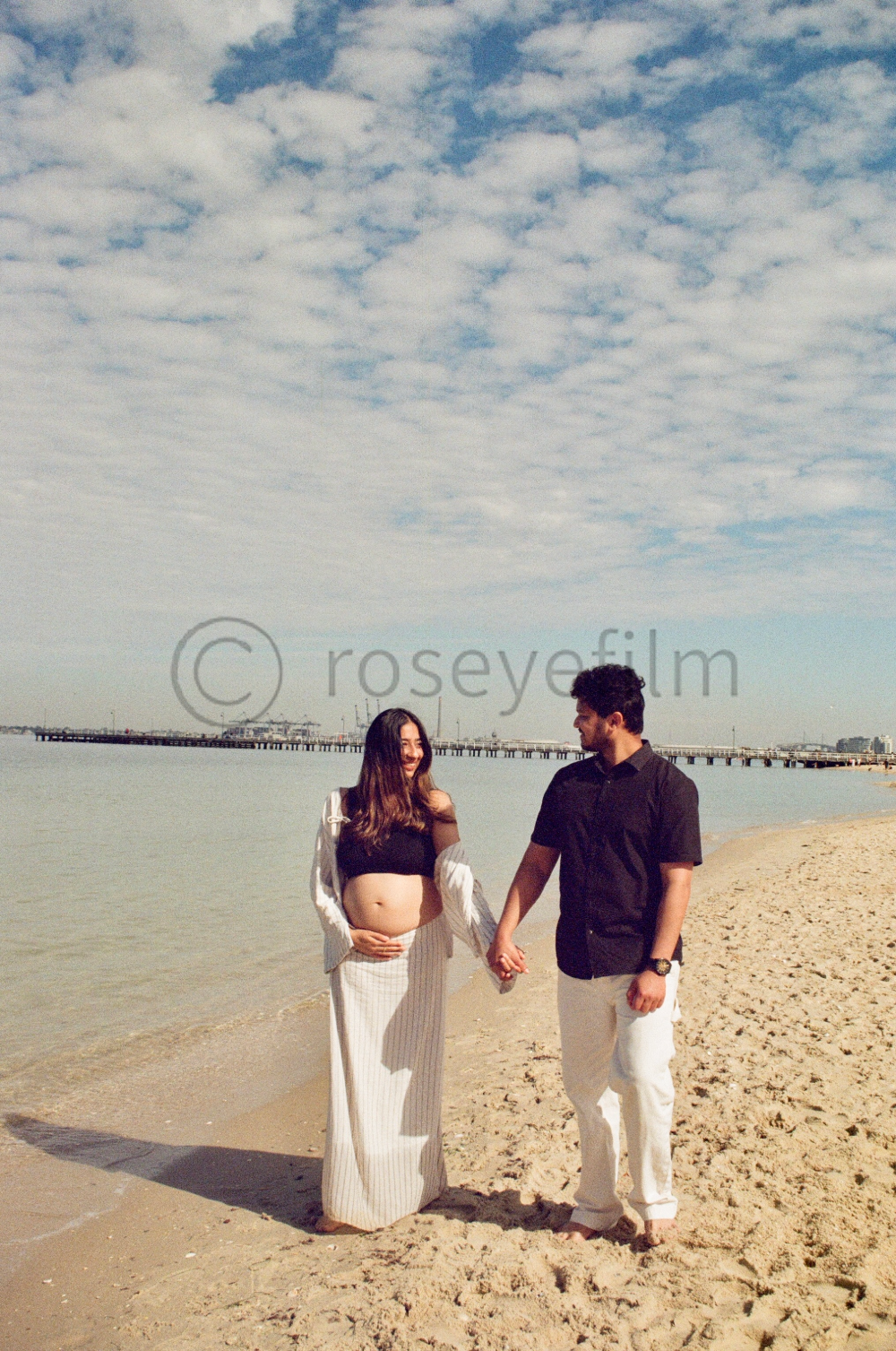 A couple holding hands on the beach, with the woman visibly pregnant, smiling and looking at each other under a partly cloudy sky.