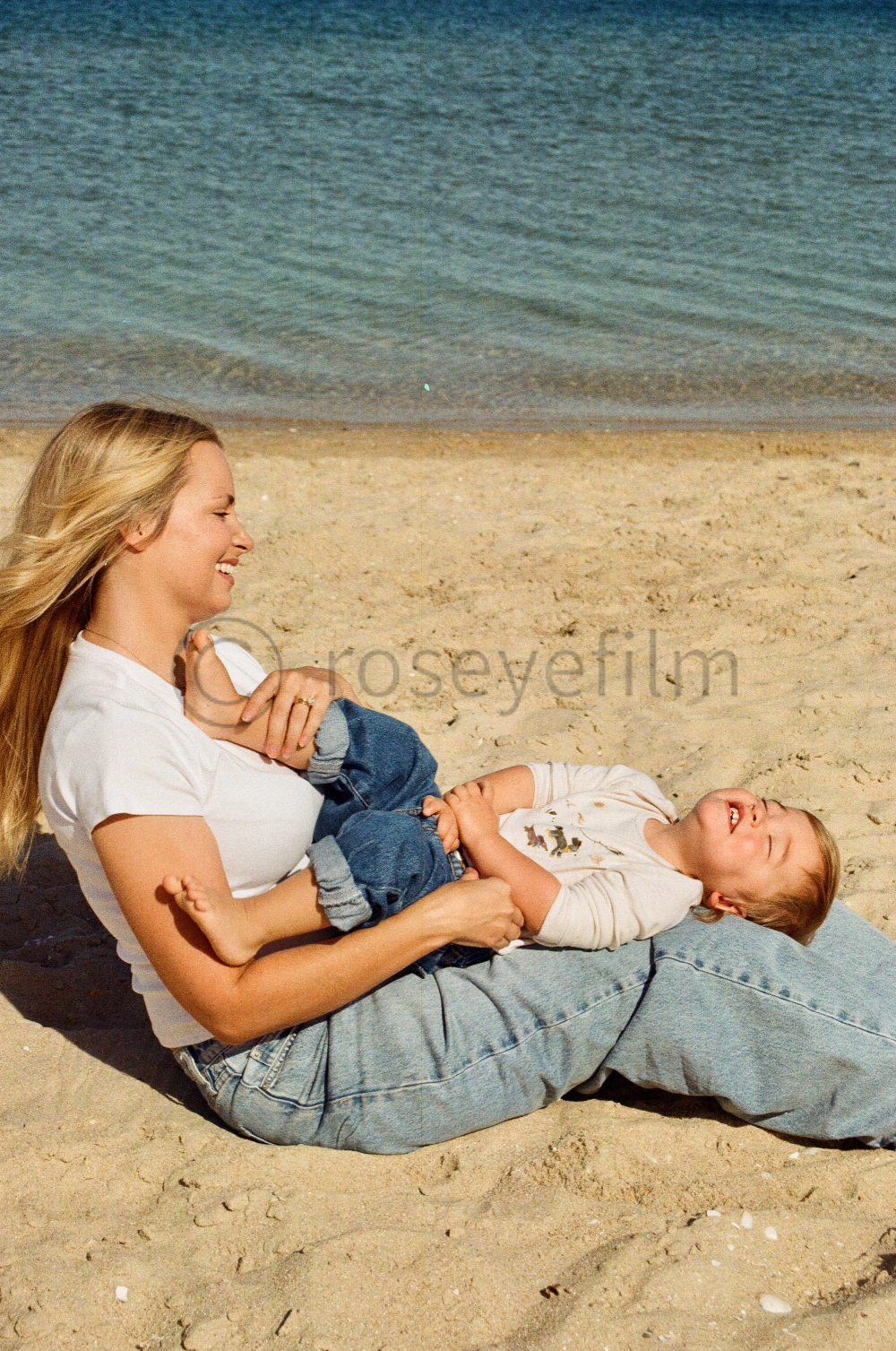 A woman and a young boy laughing and playing on a sandy beach near the water.