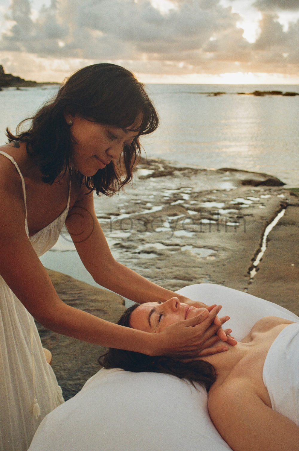 A woman is receiving a facial massage outdoors on a rocky beach during sunset from another woman who is practicing massage or skincare techniques.