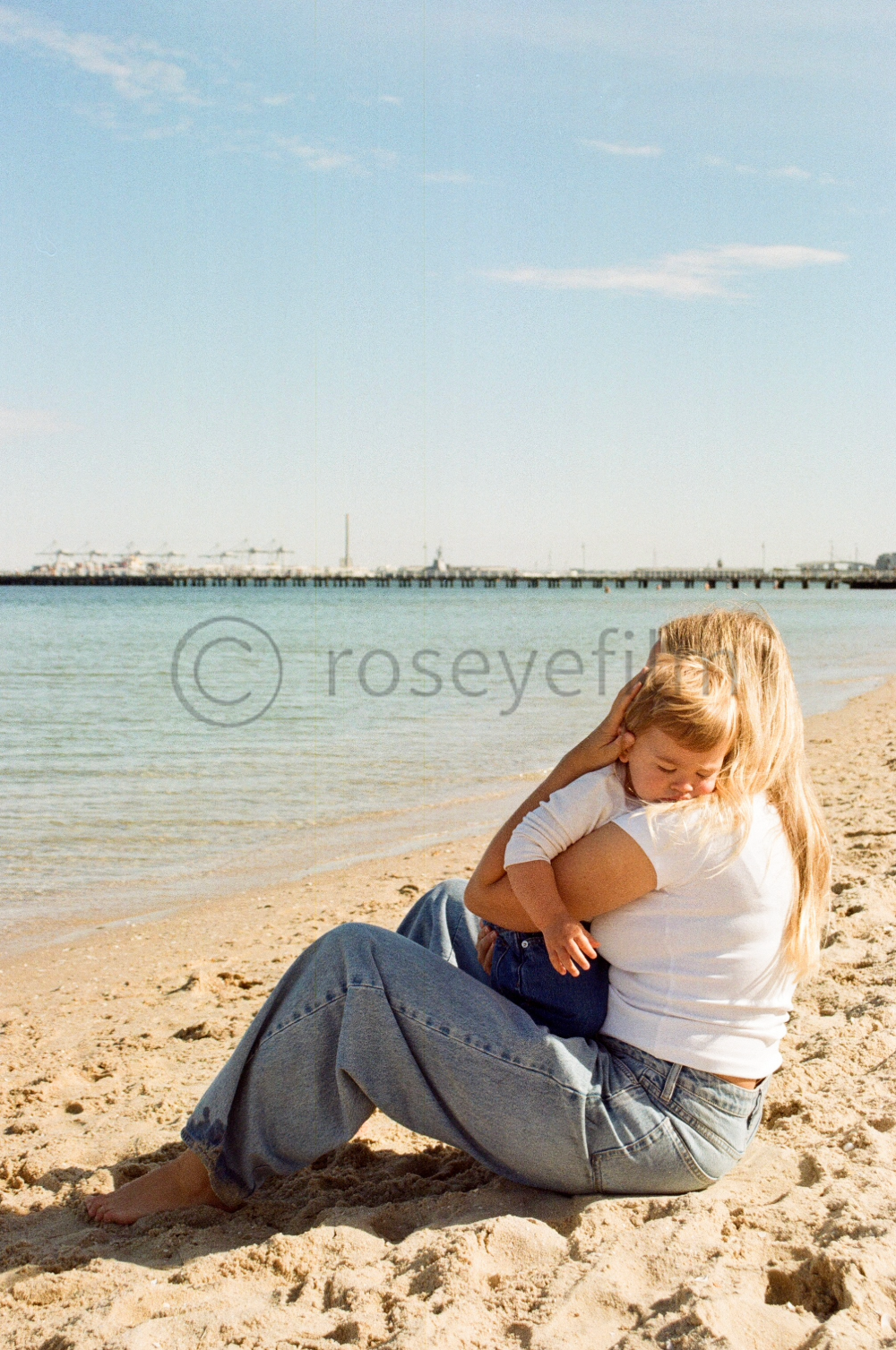 A woman sitting on the sandy beach hugging a young child, with the ocean and a pier in the background.