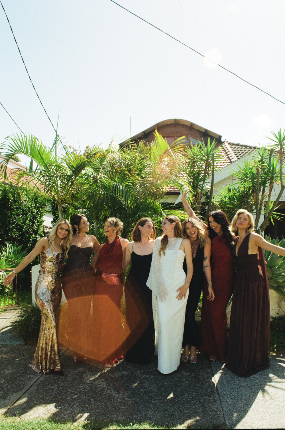 A group of women dressed in elegant evening gowns standing outside in front of lush green plants, smiling and enjoying a sunny day.