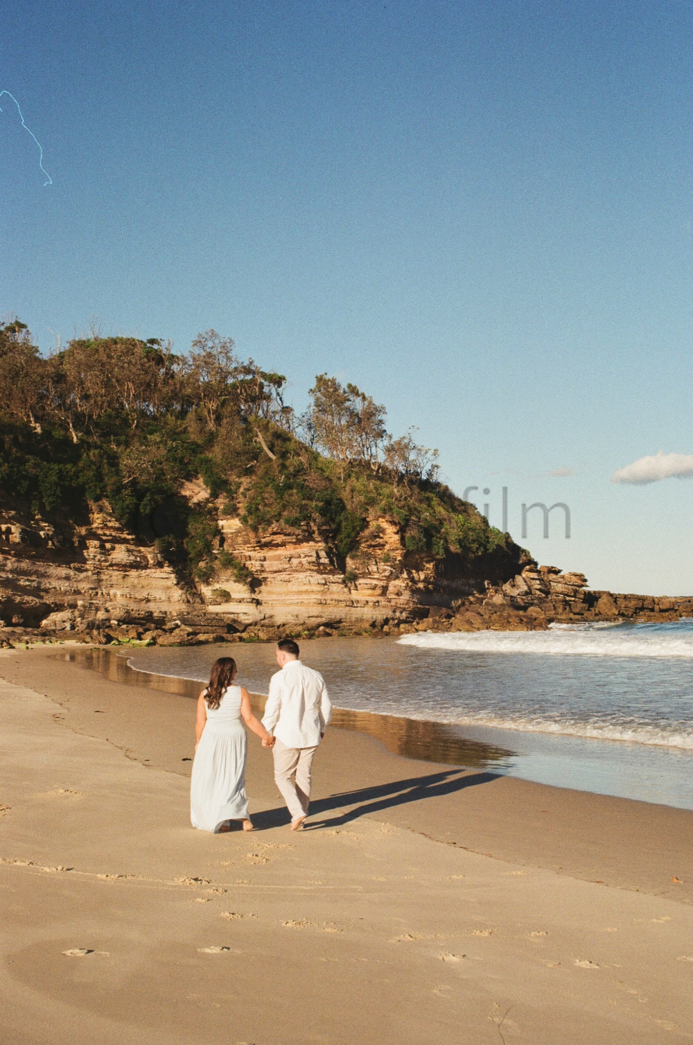 Couple walking hand in hand along the beach with cliffs and trees in the background on a clear day.